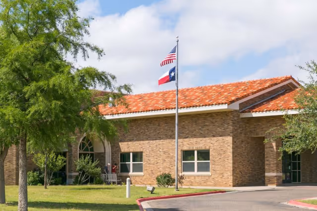 Exterior view of a single-story brick building with a red tile roof, two flagpoles displaying the American and Texas flags, surrounded by green trees and a lawn under a partly cloudy sky.