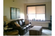 Cozy living room with brown leather sofa, ottoman, lamp, and window blinds.