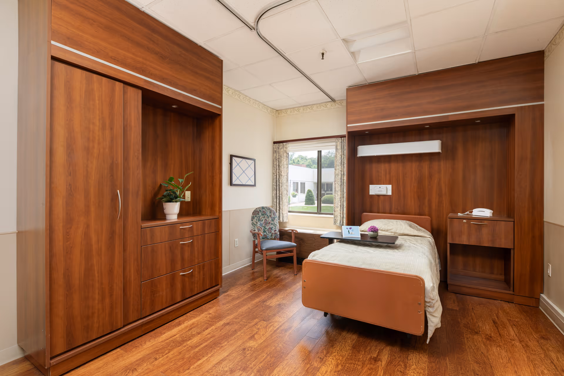 A senior living facility bedroom with a single bed featuring a beige blanket and a tray with a small plant and a card. The room has wooden furniture including a wardrobe with drawers, a bedside table with a telephone, and a chair with floral upholstery near a window with curtains. The floor is wooden, and the walls are light-colored with a decorative border near the ceiling.