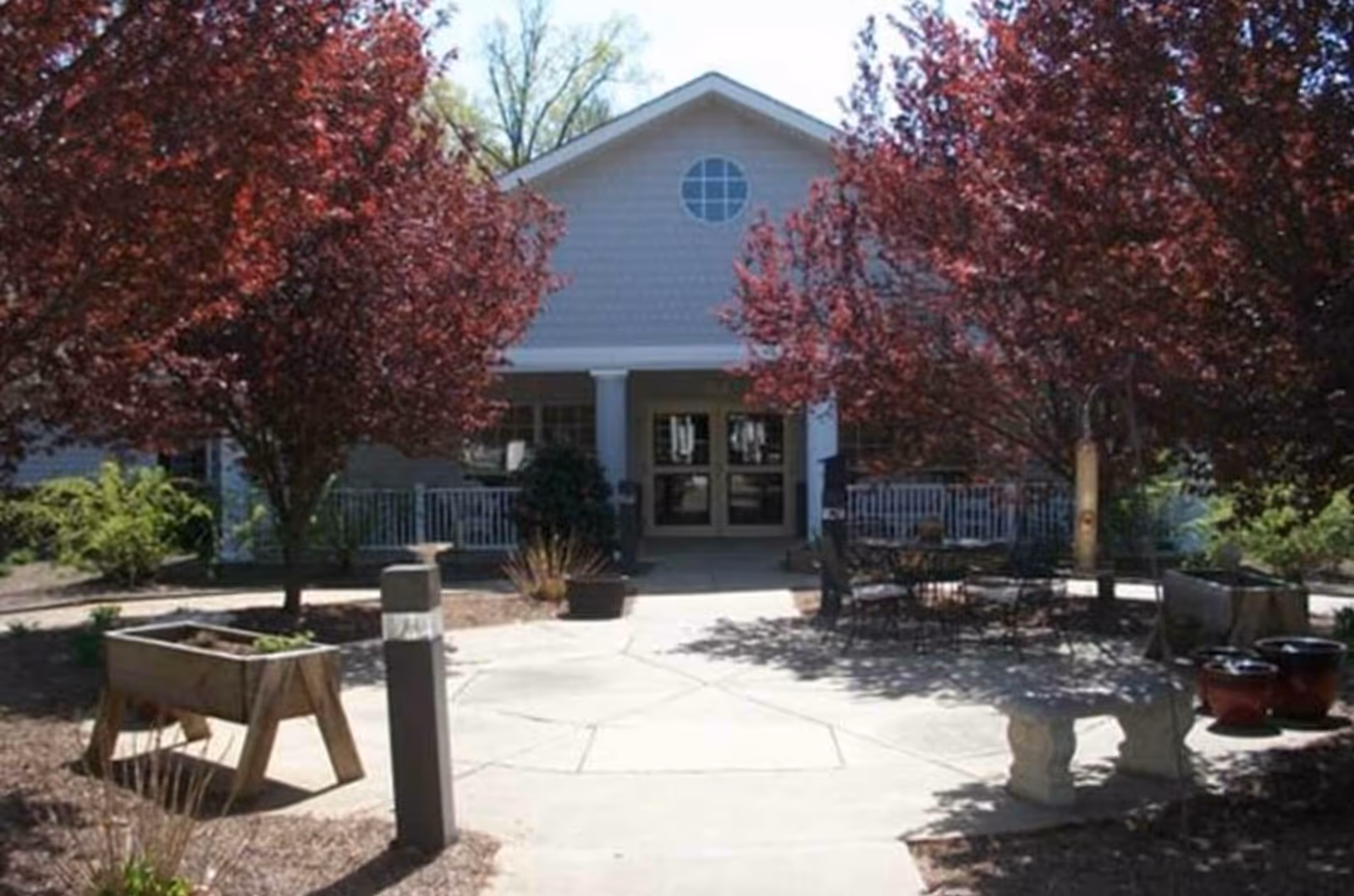 Outdoor courtyard area with a concrete pathway leading to the entrance of a building. The area is landscaped with trees that have reddish leaves, potted plants, a wooden planter box, outdoor seating with a table and chairs, and a stone bench.