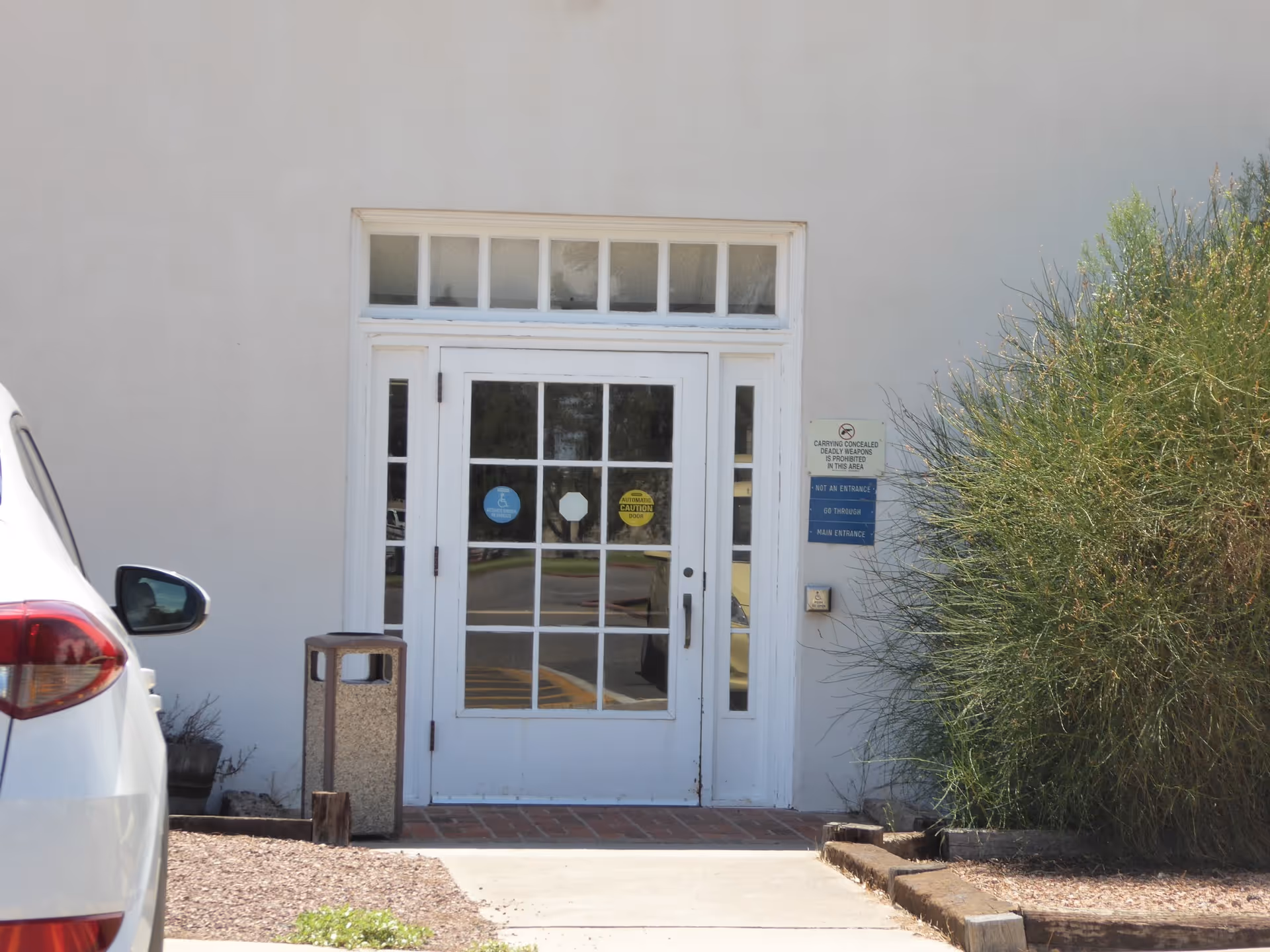 Front entrance of New Mexico State Veterans Home with white doors, parked car, and shrubs.