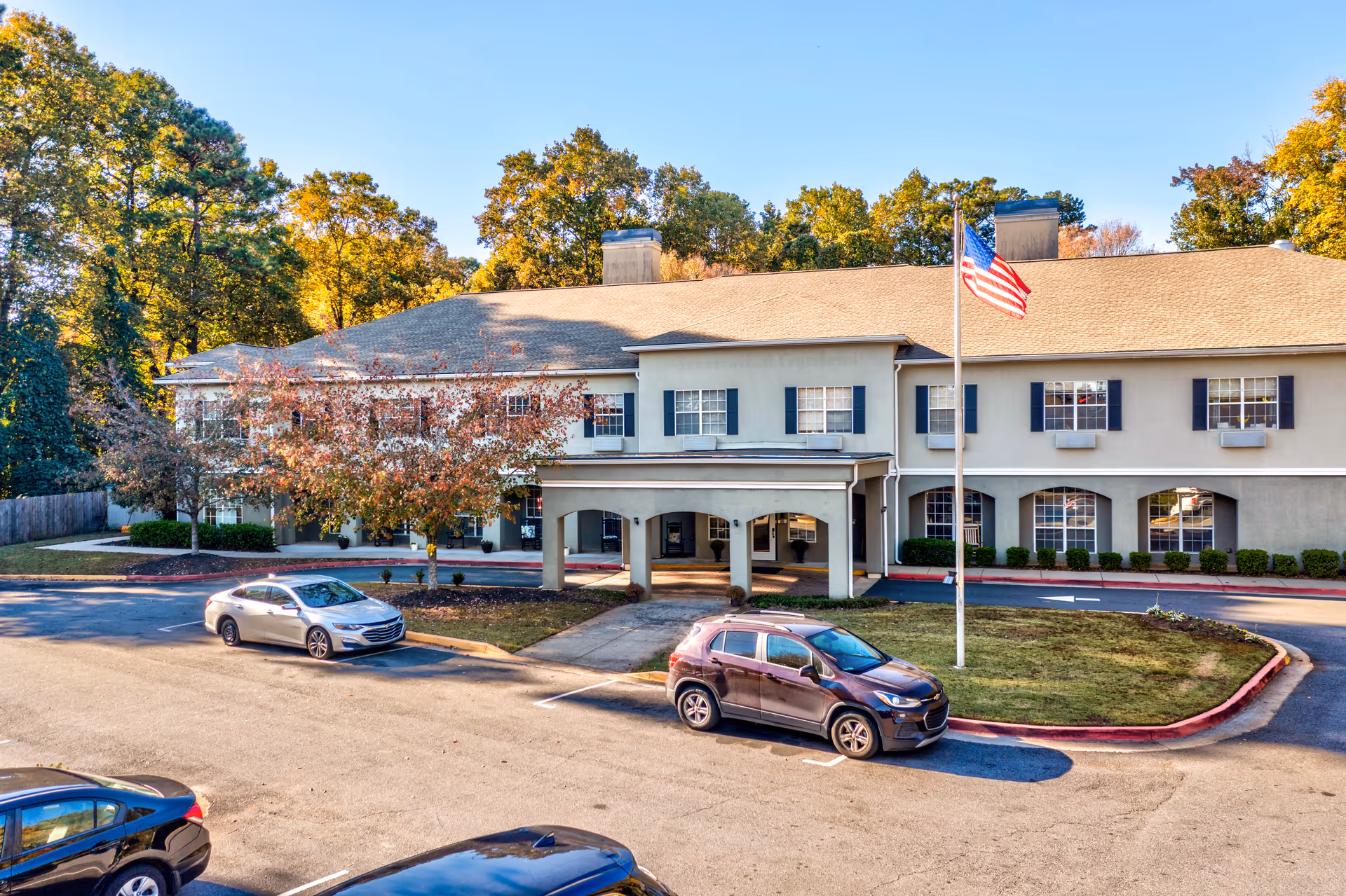 Front exterior of a two-story assisted living building with a covered entrance, an American flag, parked cars, and surrounding trees.