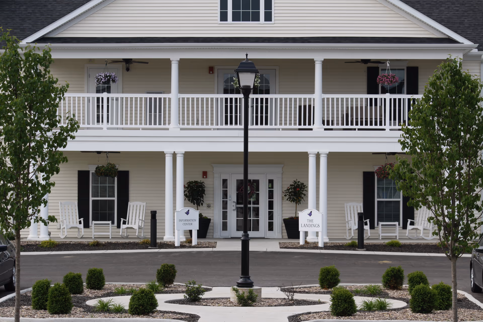 Front exterior view of a two-story senior living community building with white railings and columns, rocking chairs on the porch, hanging flower baskets, and a central lamp post in a landscaped area with small bushes and trees.