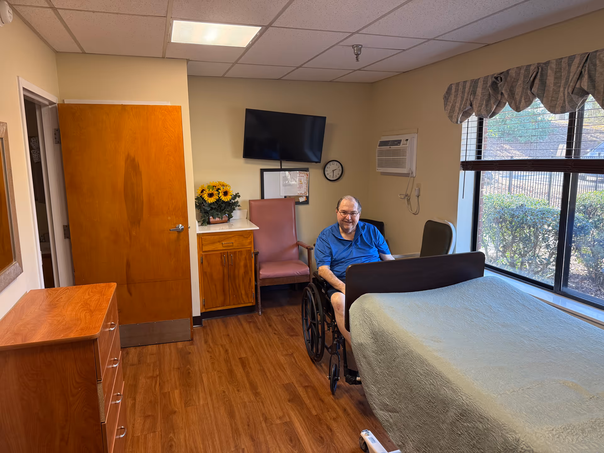 Resident in wheelchair sits beside a bed in a patient room.