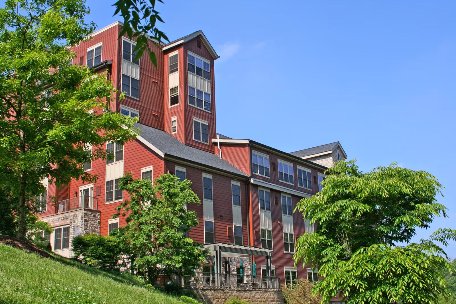 Exterior view of a multi-story senior living facility building with red siding and multiple windows, surrounded by green trees and grass under a clear blue sky.