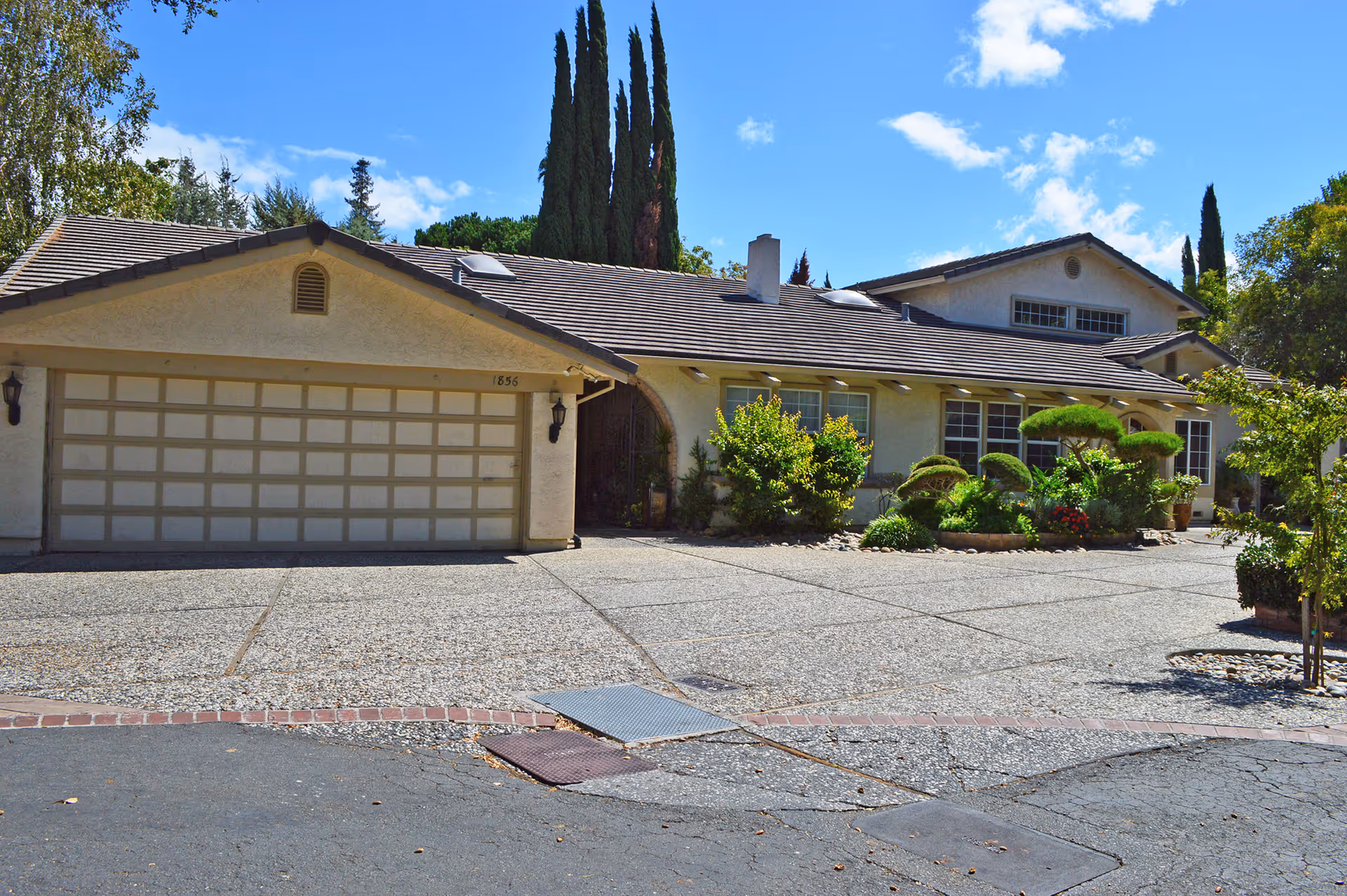 Exterior view of a single-story residential building with a large garage door, surrounded by neatly trimmed bushes and trees under a blue sky with some clouds.