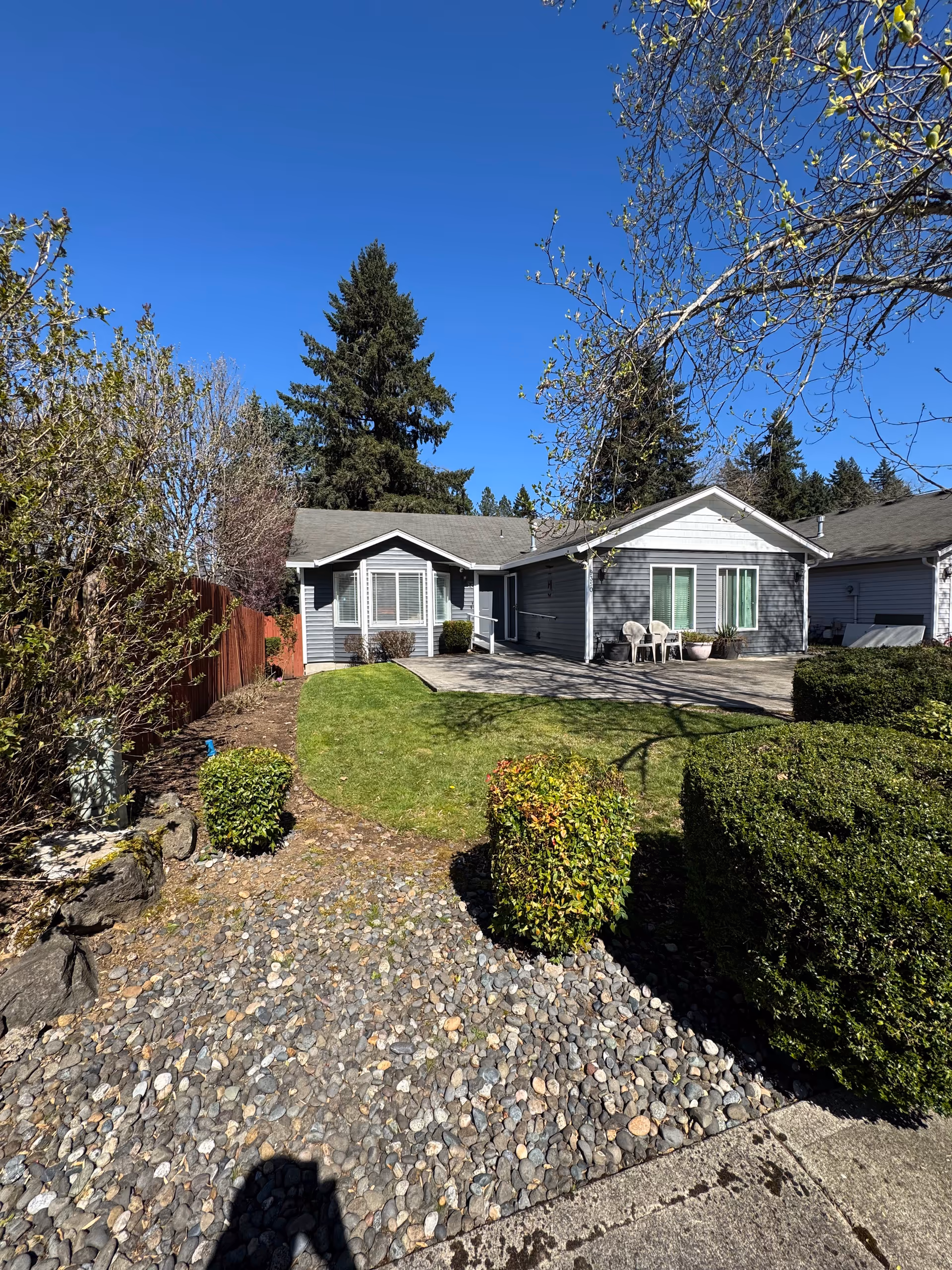 Single-story gray house with a front yard, shrubs, and outdoor seating on a sunny day.
