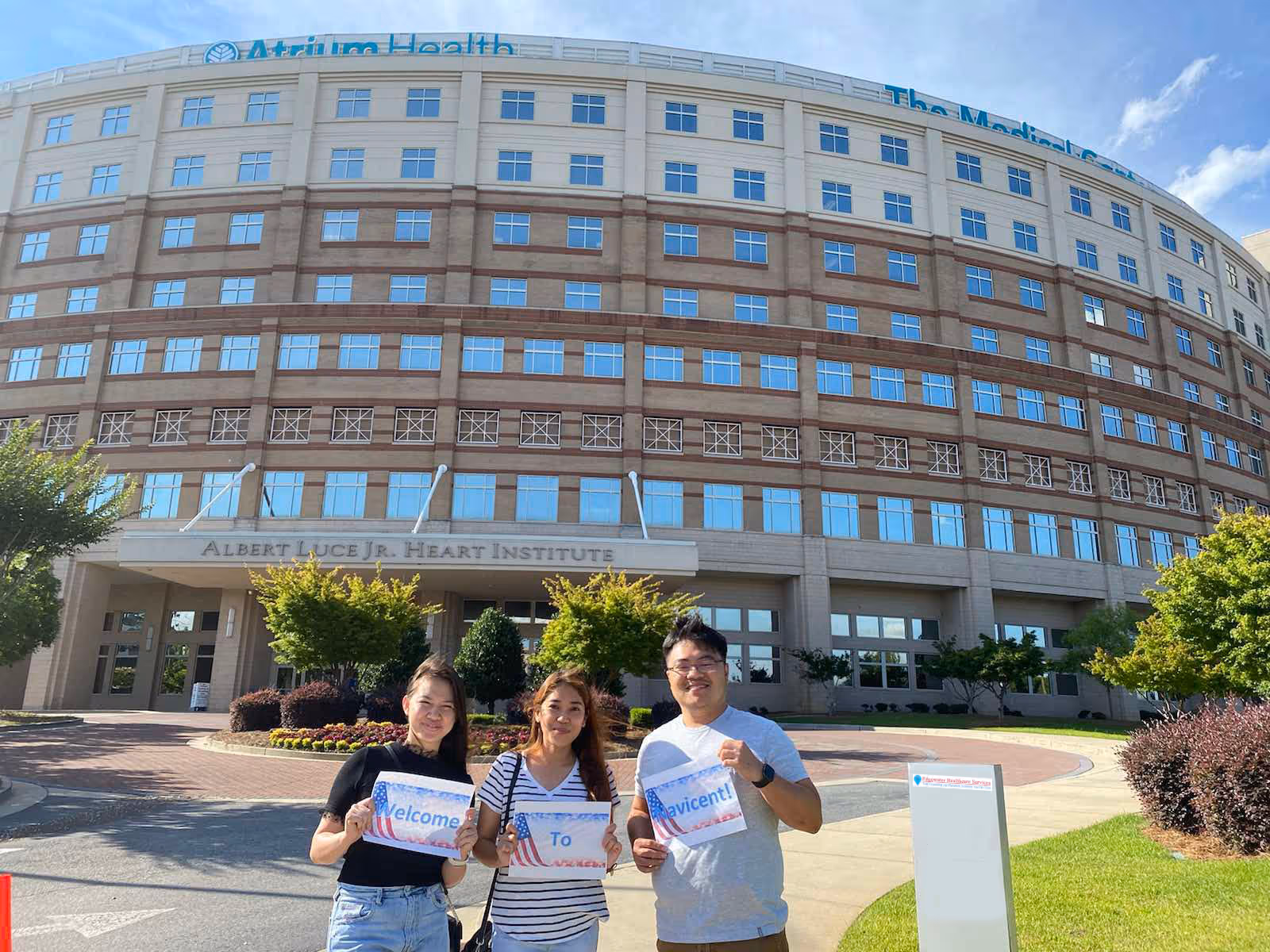 Three people stand smiling in front of a large hospital building with signage.