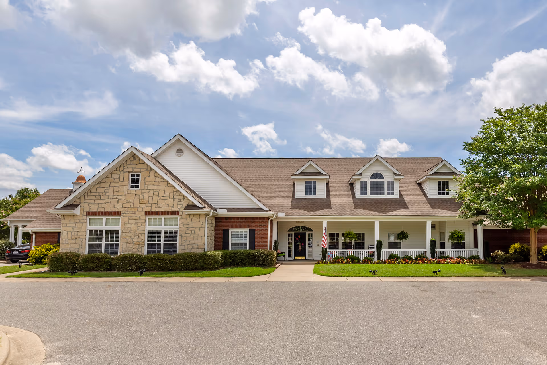 Front exterior view of a single-story senior living facility building with a combination of stone and brick facade, multiple windows, a covered porch with white railings, hanging plants, and a well-maintained lawn under a partly cloudy sky.