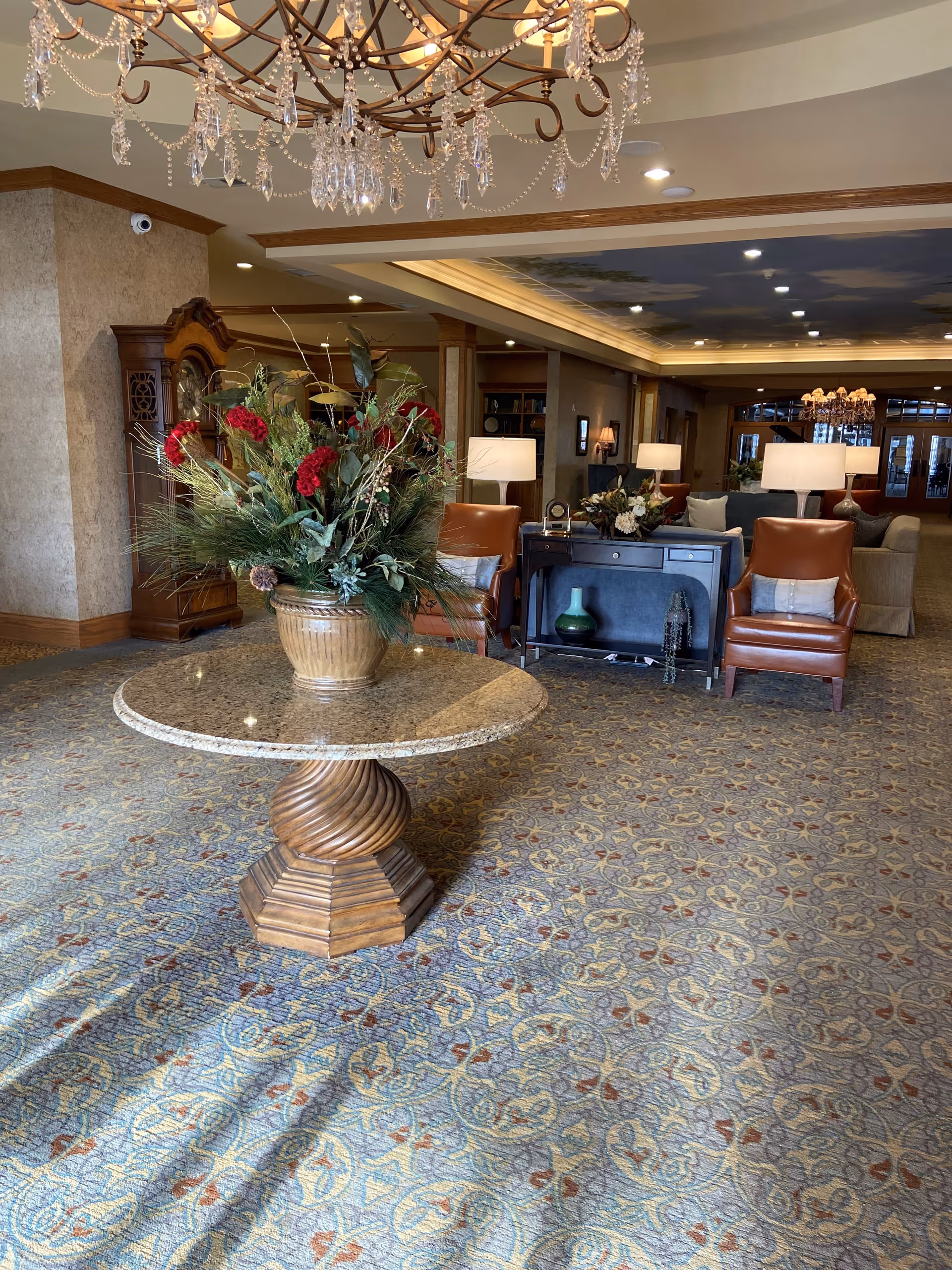 Elegant senior-living lobby with round granite table, floral arrangement, chandelier, and seating.