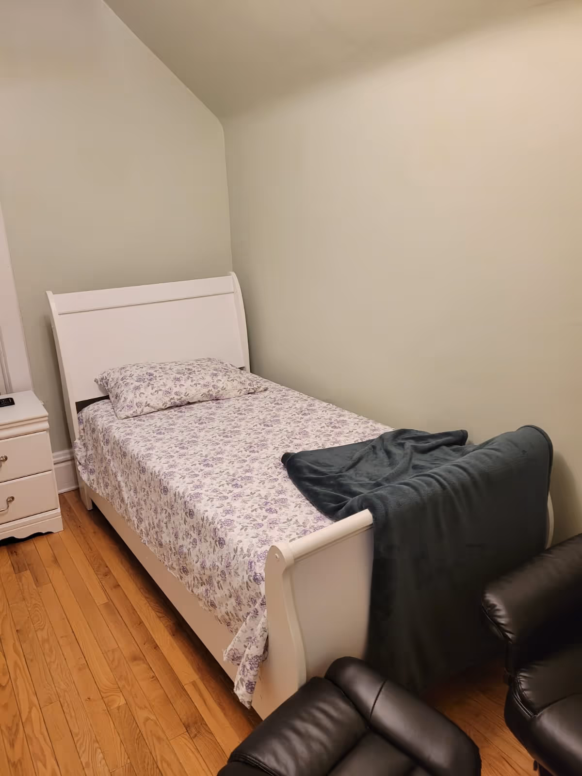 Small white bedroom with single bed, floral sheets, and a dark leather chair.