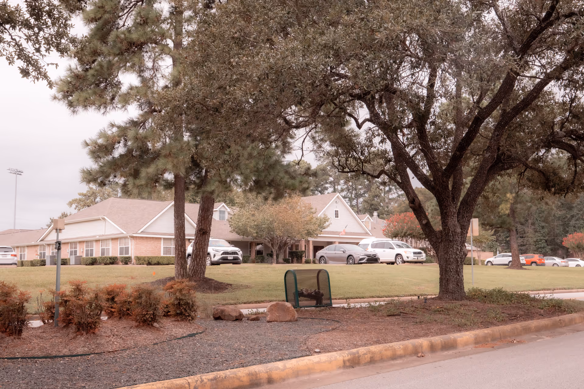Front exterior of a single-story senior living facility with lawn, trees, and several parked cars.