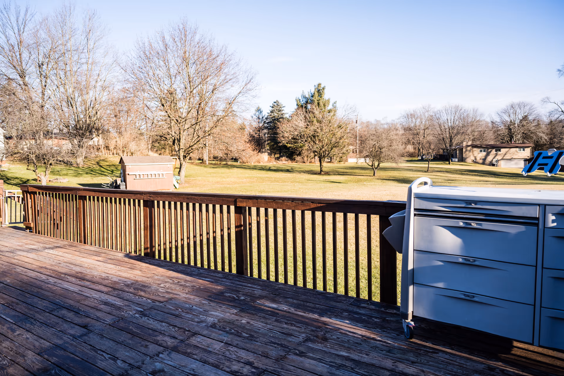 View from a wooden deck overlooking a large grassy yard with scattered leafless trees and a small shed. There is a gray storage cabinet with drawers on the right side of the deck. The sky is clear and blue.