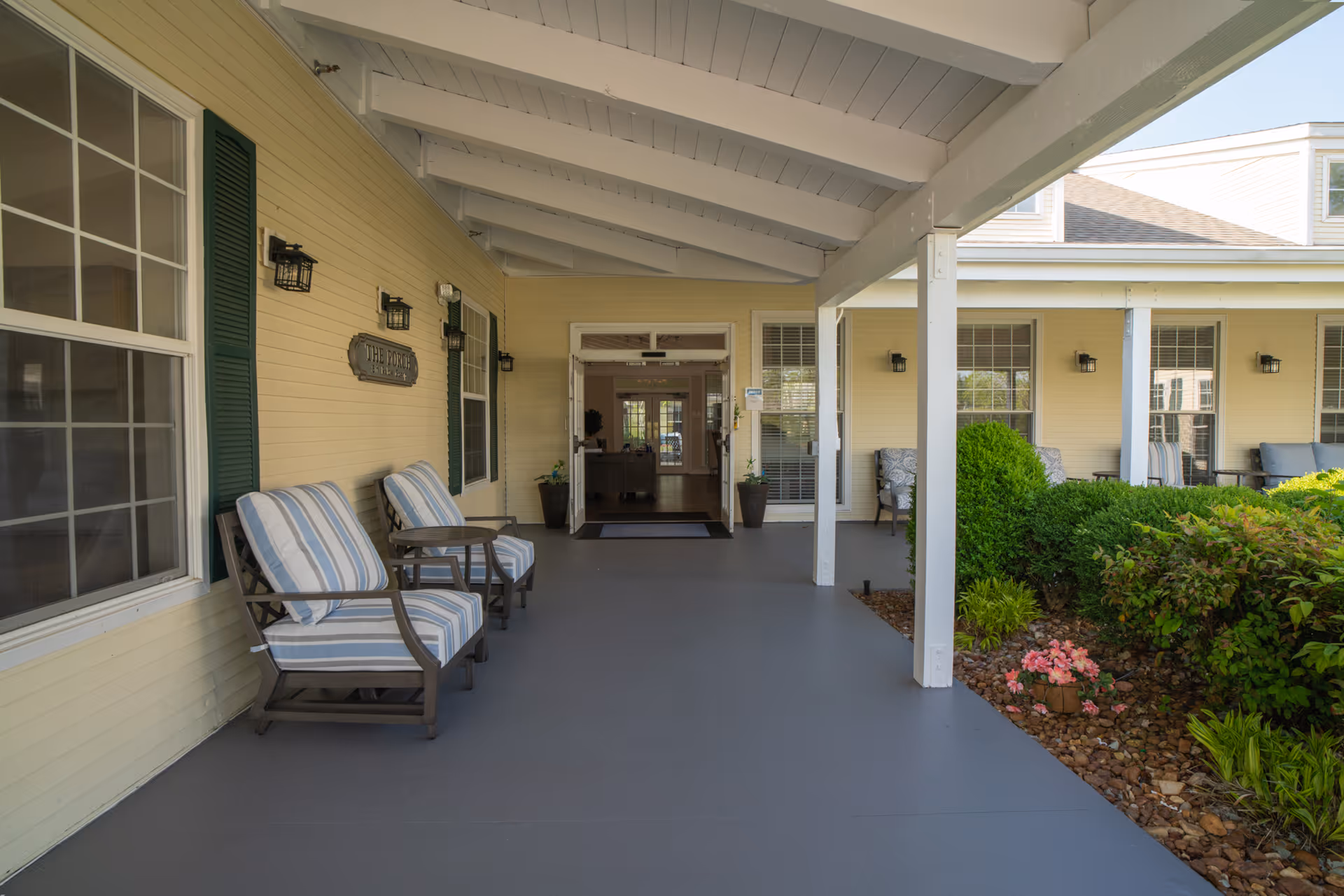 Covered front porch entrance with striped cushioned chairs, columns, and landscaping leading to double doors.
