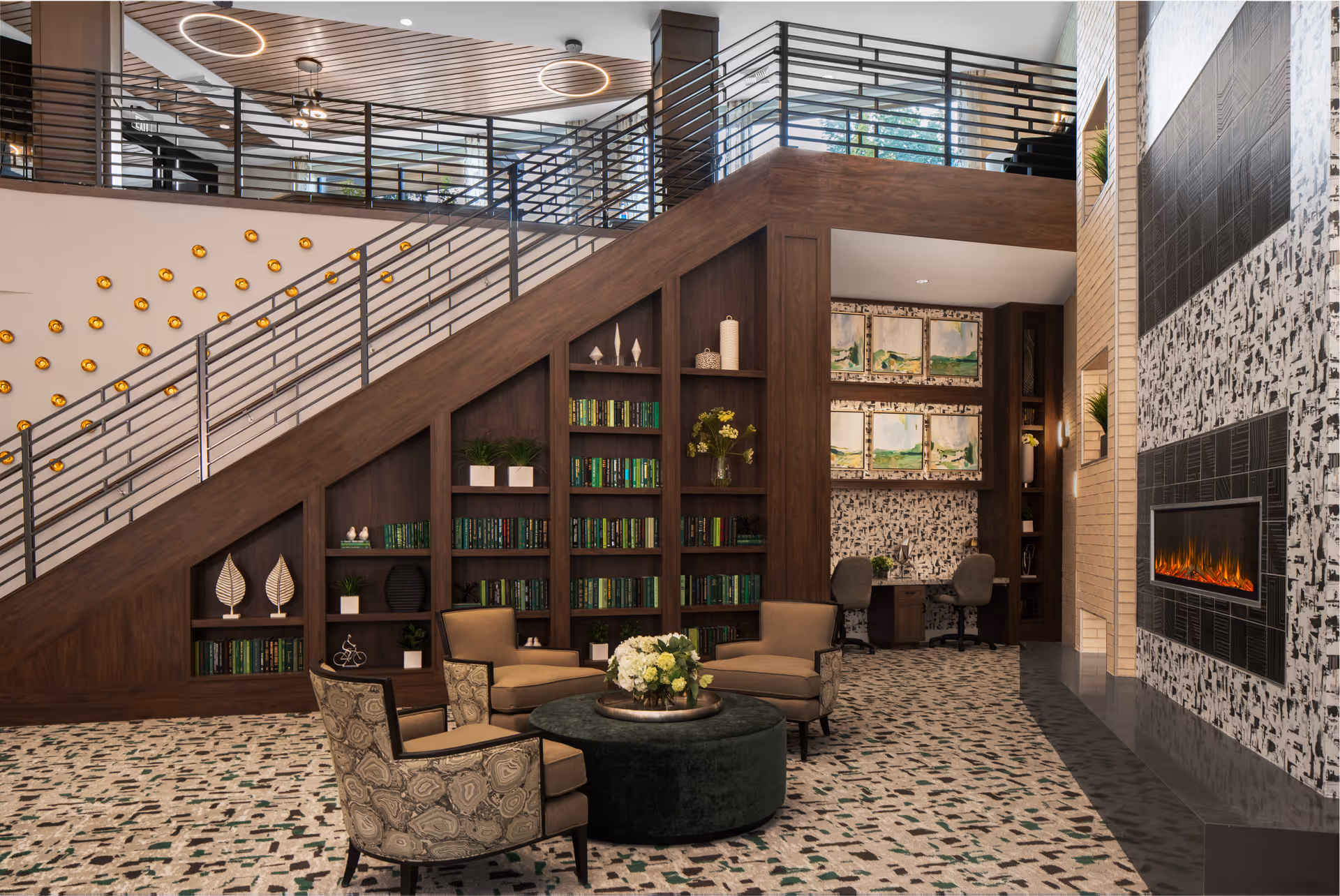 A modern and cozy living room area in an assisted living facility featuring a built-in wooden bookshelf under a staircase filled with books and decorative items. There are four upholstered chairs arranged around a round ottoman with a flower arrangement on top. The room has a patterned carpet, a wall-mounted electric fireplace with a black and white abstract design above it, and a small workspace with two chairs and framed artwork on the wall in the background. The upper level is visible with metal railings and circular ceiling lights.