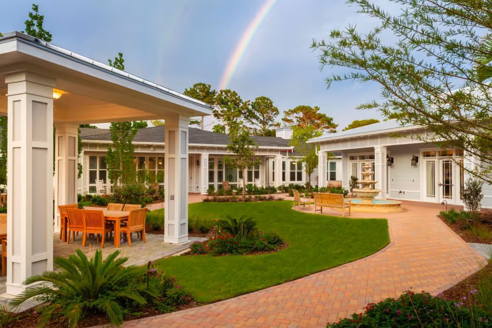 Outdoor courtyard area at Mount Pleasant Gardens featuring a covered seating area with wooden chairs and tables, a brick pathway, green lawn, plants, trees, a water fountain, and a double rainbow in the sky.