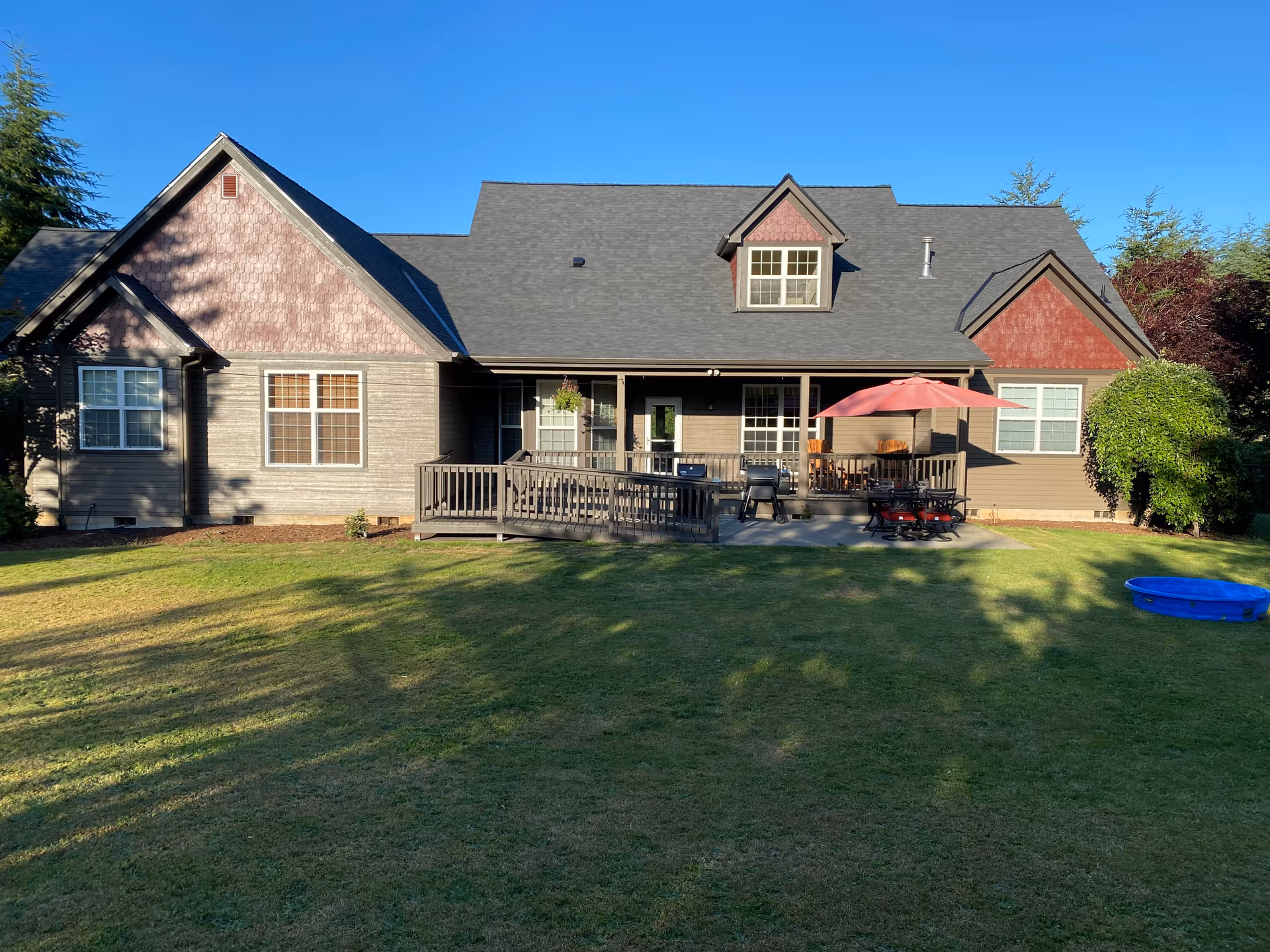 Single-story house with a wooden deck, outdoor furniture, red umbrella, and expansive green lawn.