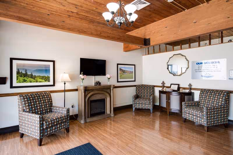 Communal living room with patterned armchairs, an electric fireplace below a wall-mounted TV, framed artwork, and a wooden ceiling chandelier.