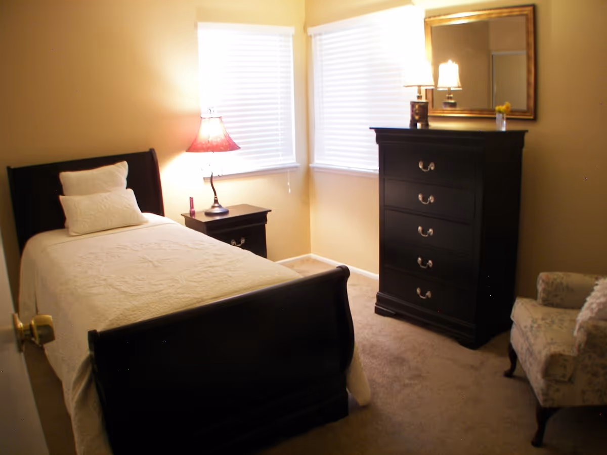 A cozy bedroom with a single bed, nightstand, dresser, mirror, and upholstered chair.