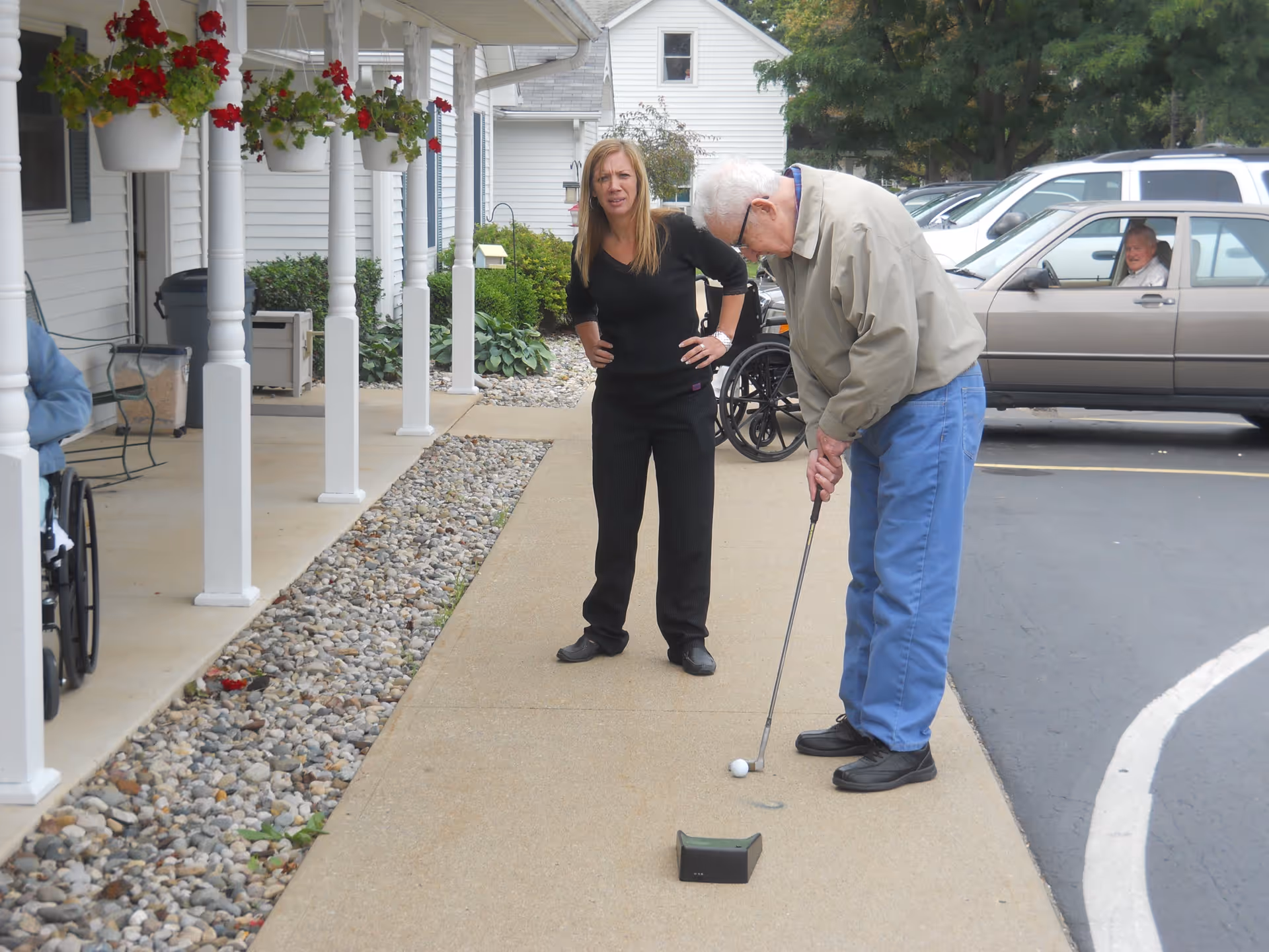 An elderly man is playing mini golf on a concrete pathway outside a building, with a woman standing nearby watching him. There are hanging flower pots with red flowers along the porch, a wheelchair visible to the left, and parked cars in the background.