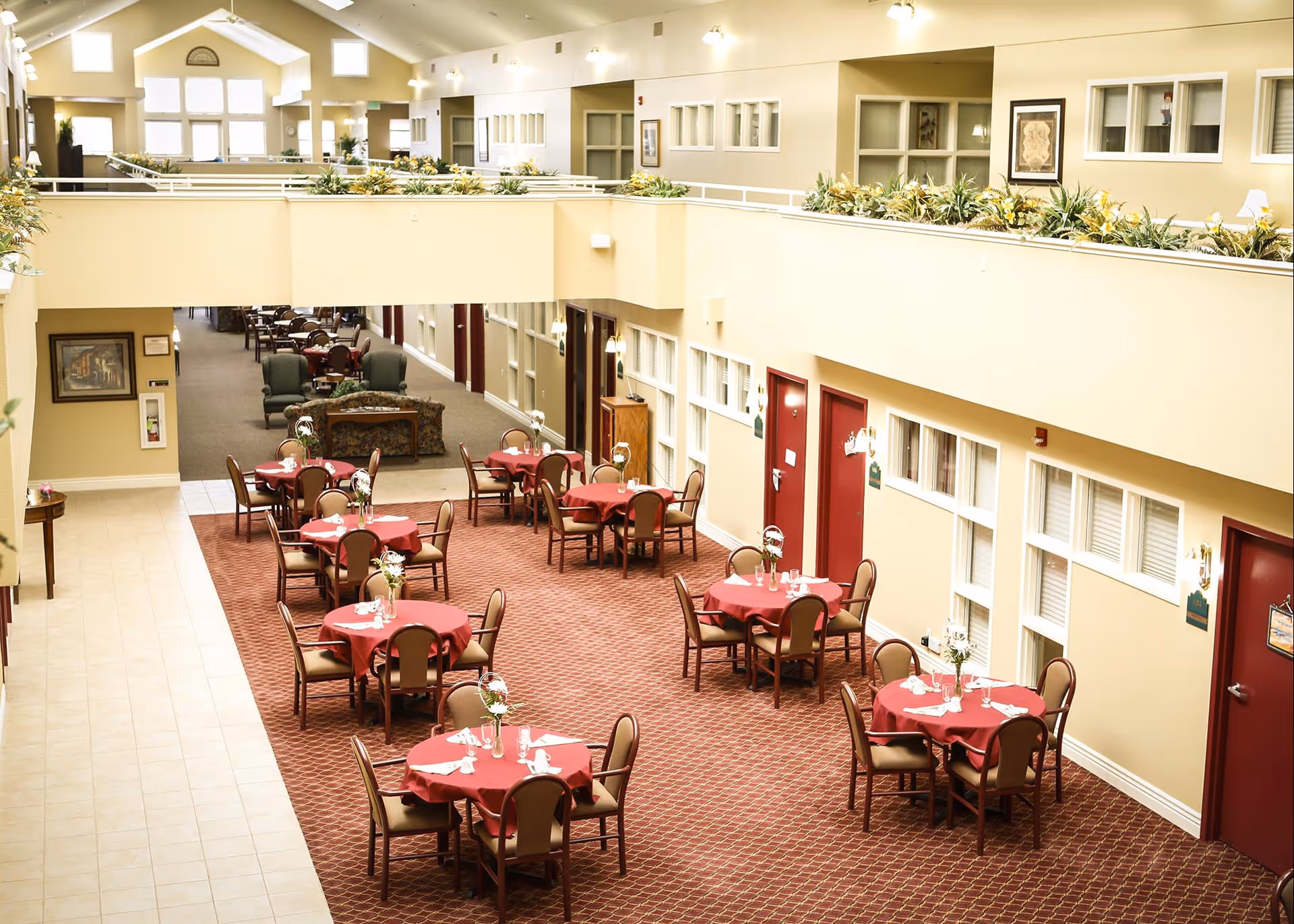 Interior view of a dining area in an assisted living facility with multiple round tables covered with red tablecloths, each set with napkins and small flower arrangements. The space has beige walls, red doors, and a carpeted floor. There is a second-floor balcony with plants along the railing and additional seating visible in the background.