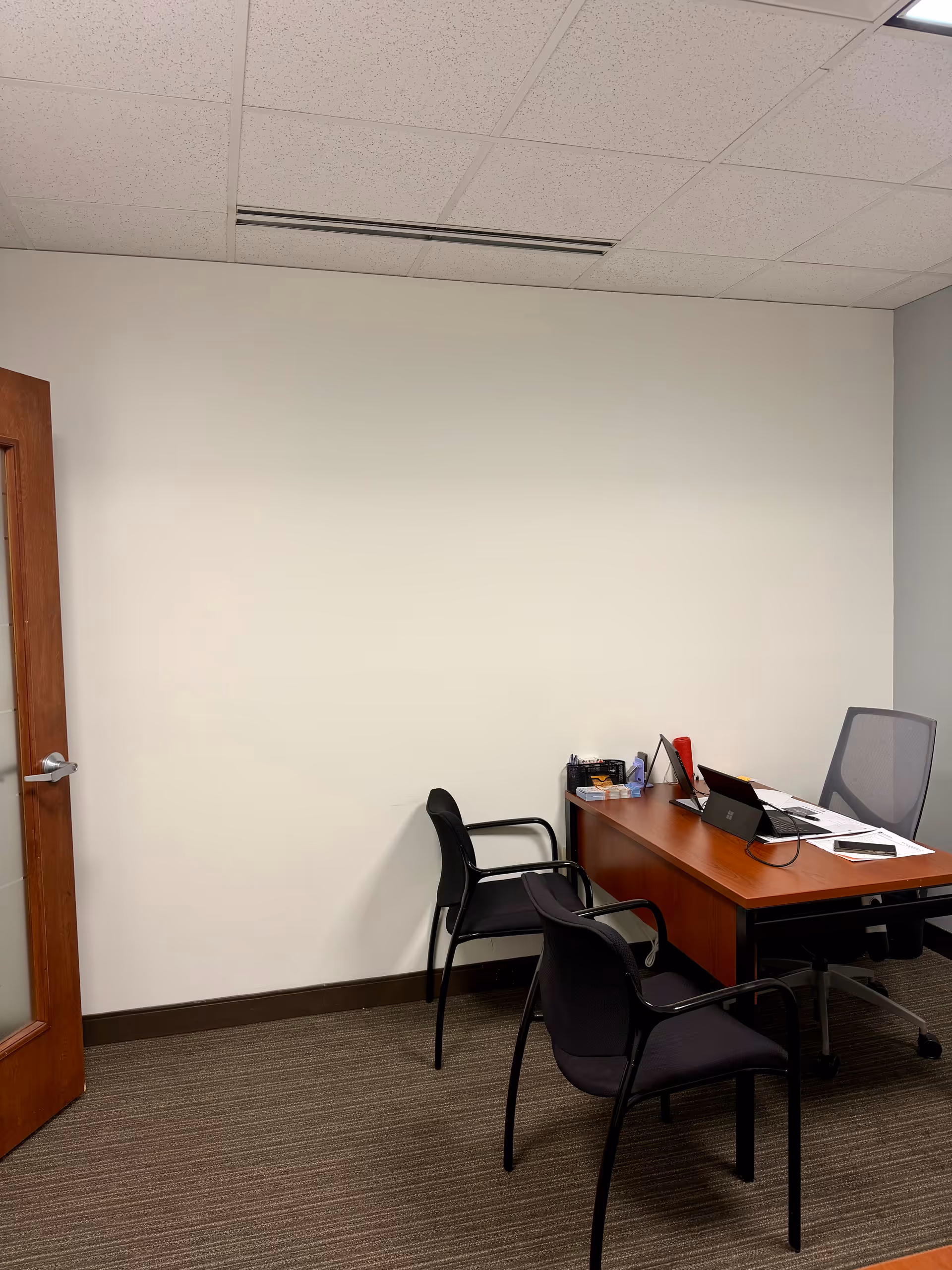A small office with a wooden desk, chairs, laptop, and scattered paperwork.