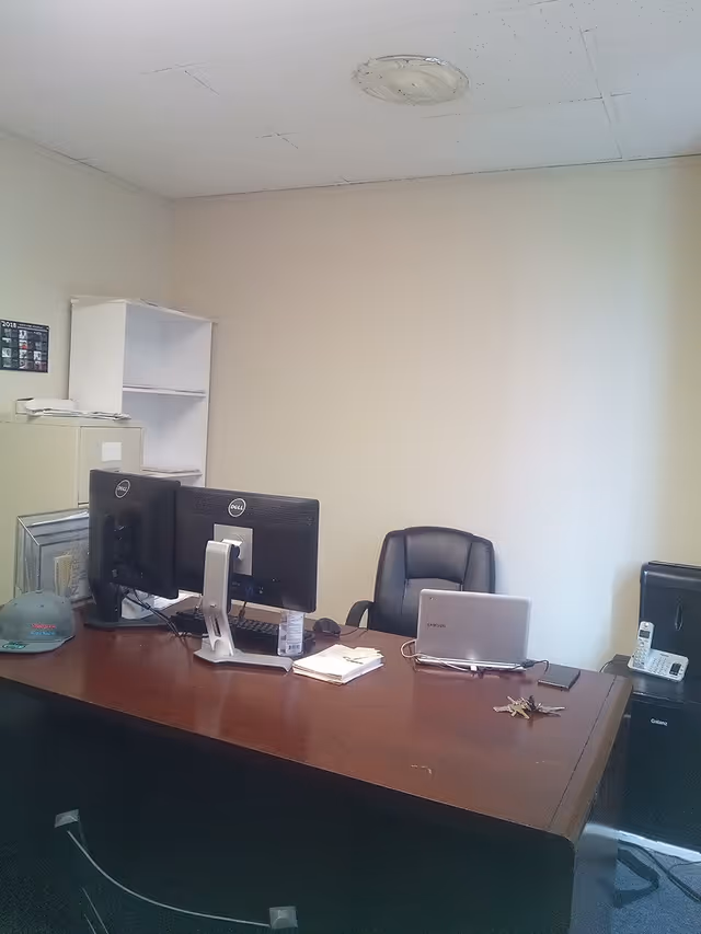 Facility office with desk, two monitors, laptop, and scattered papers.