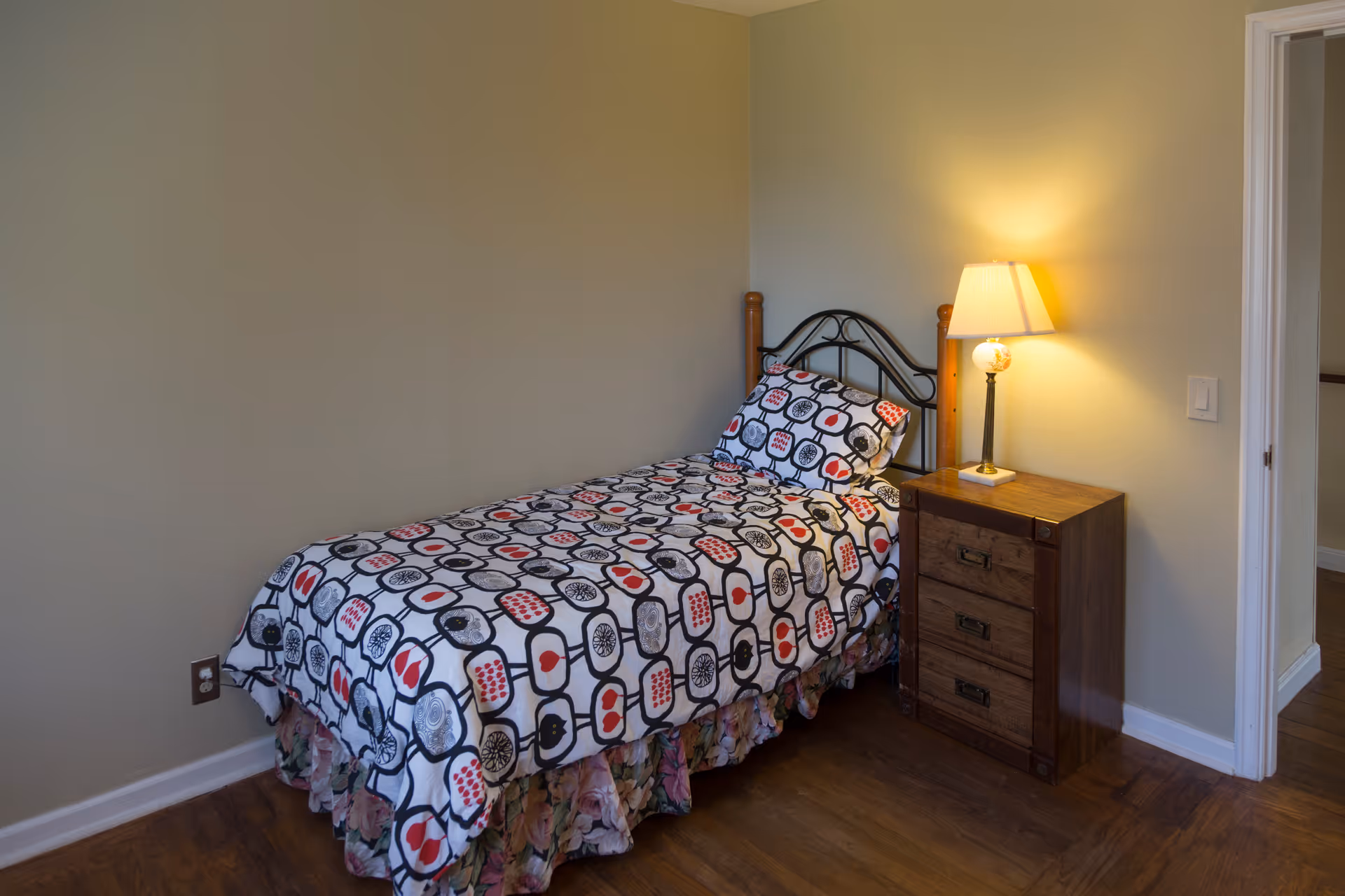 A small bedroom with a single bed covered in a patterned bedspread featuring black, white, and red designs. Next to the bed is a wooden nightstand with three drawers and a lit table lamp. The walls are painted light beige, and the floor is wooden. There is an open doorway visible on the right side of the image.