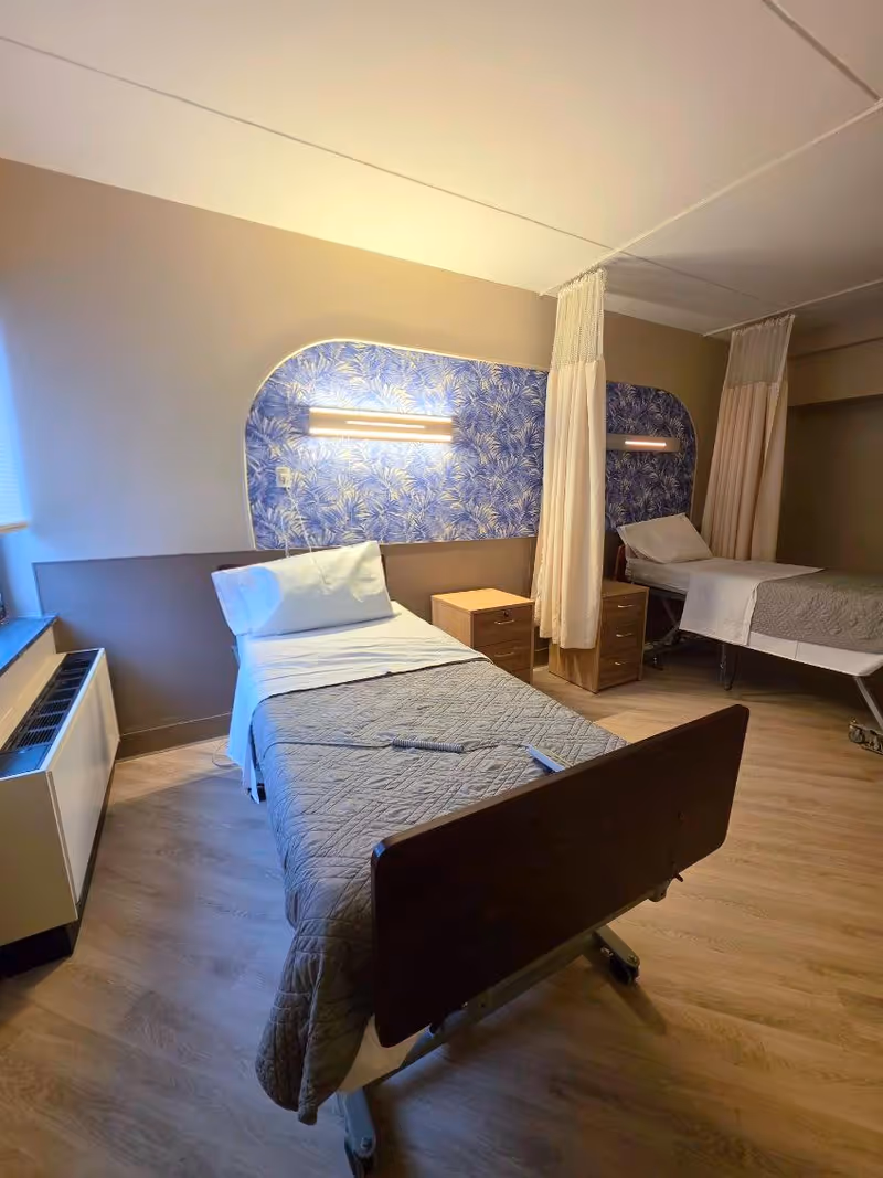 Two hospital-style beds with curtains, nightstands, and blue patterned headboard in a shared patient room.
