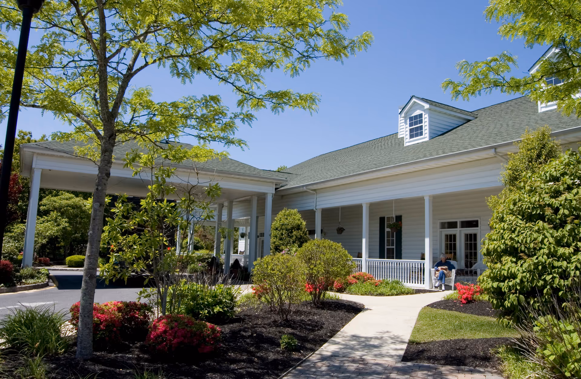 Exterior view of a senior living facility with a covered entrance, white siding, green roof, and a porch with seating. The surrounding area features landscaped bushes, trees, and a paved walkway under a clear blue sky.