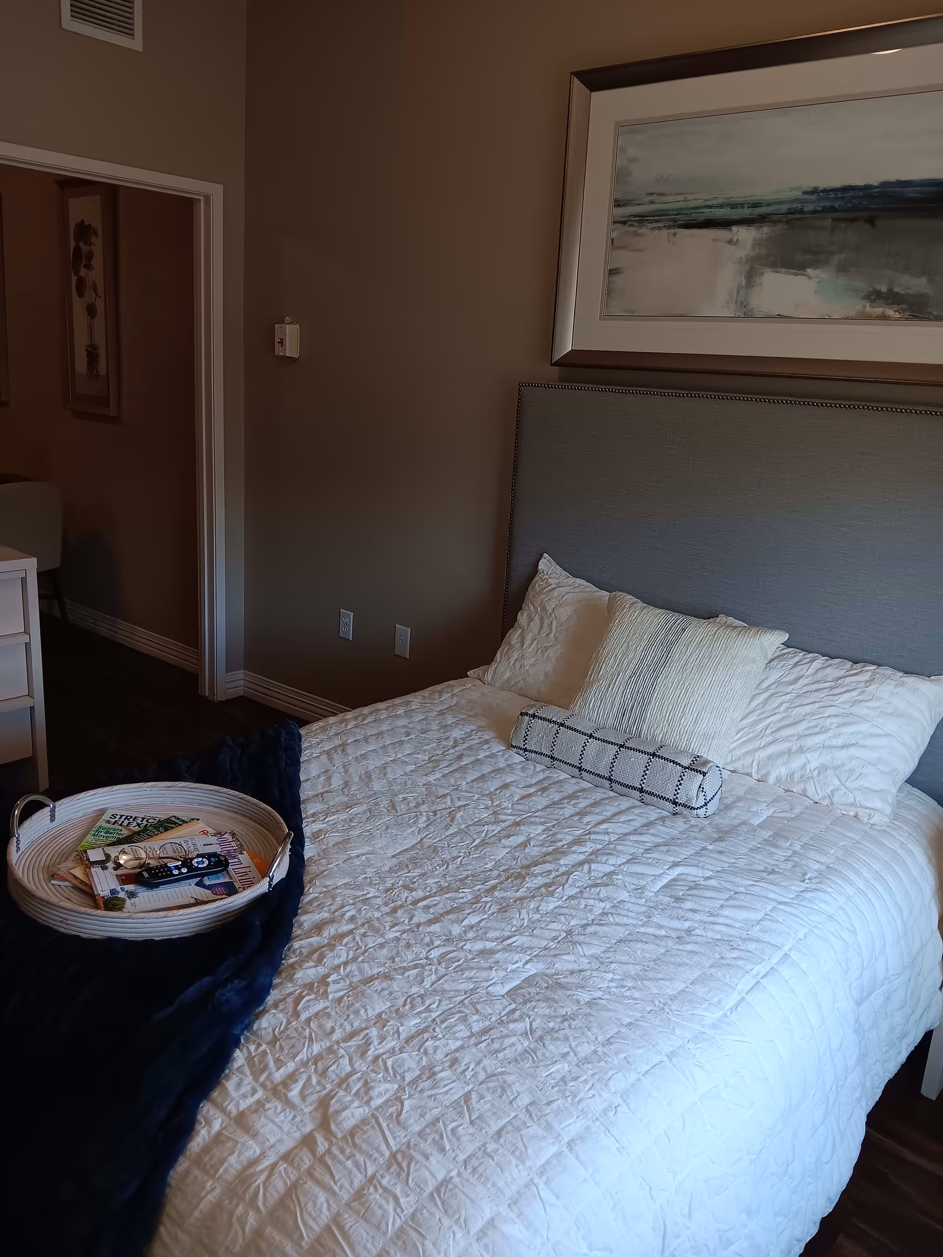 A neatly made bedroom with a white quilted bed, decorative pillows, a gray upholstered headboard, and a tray with magazines on the bed.