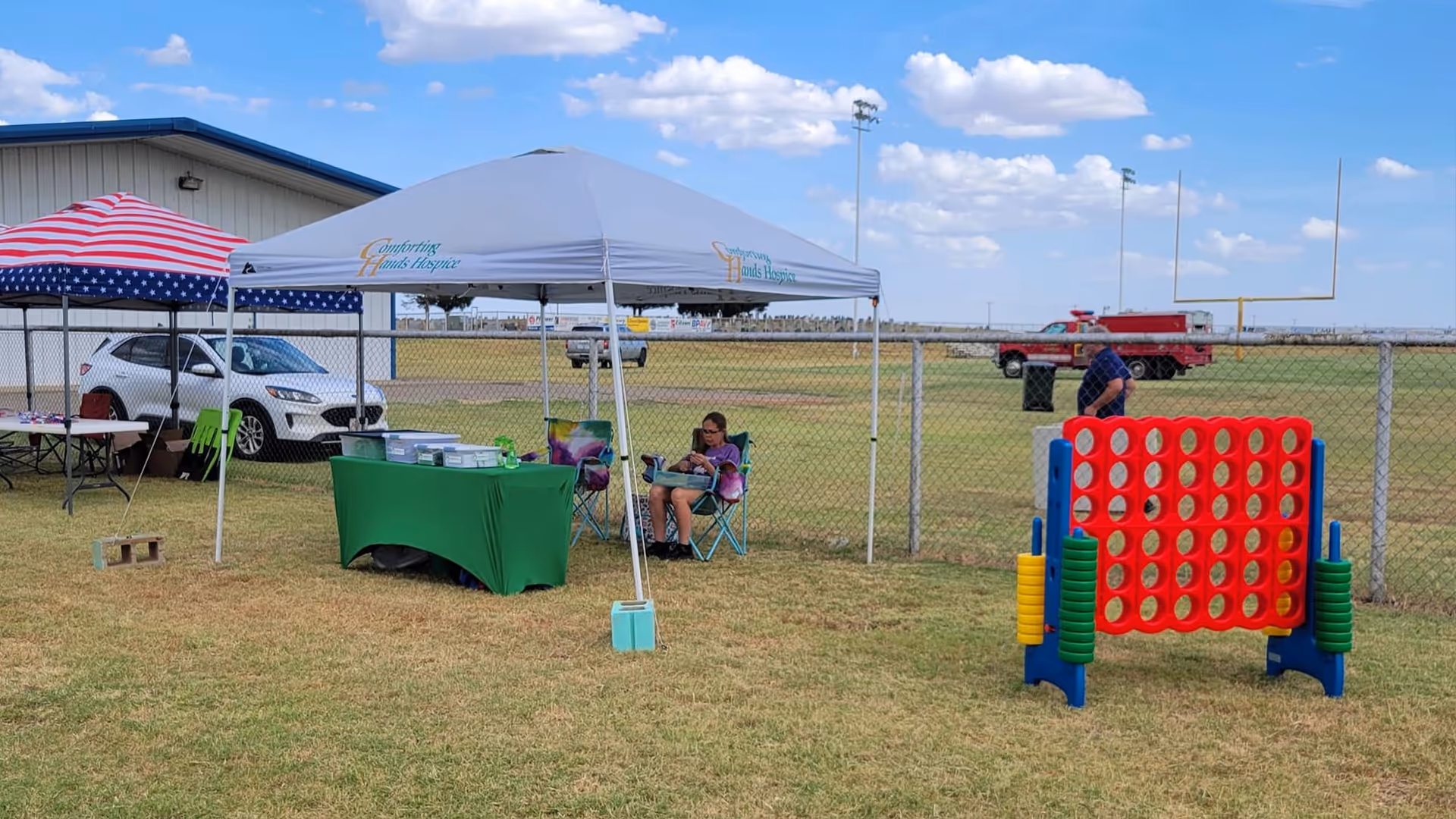Outdoor event area with tents, a green table, people sitting, and a large Connect Four game.