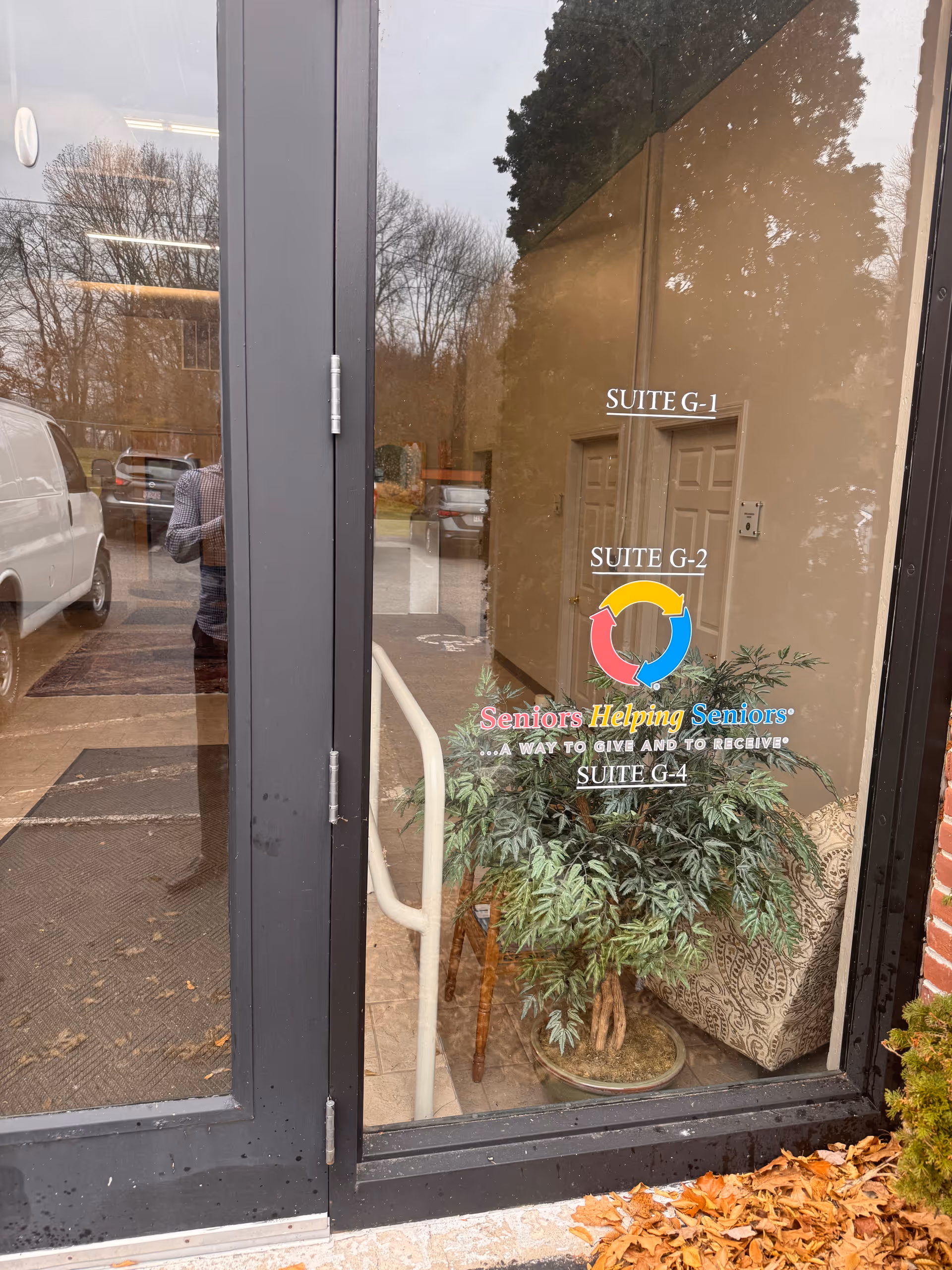 Glass entrance door displays suite signs and a colorful Seniors Helping Seniors logo, with outside reflections.