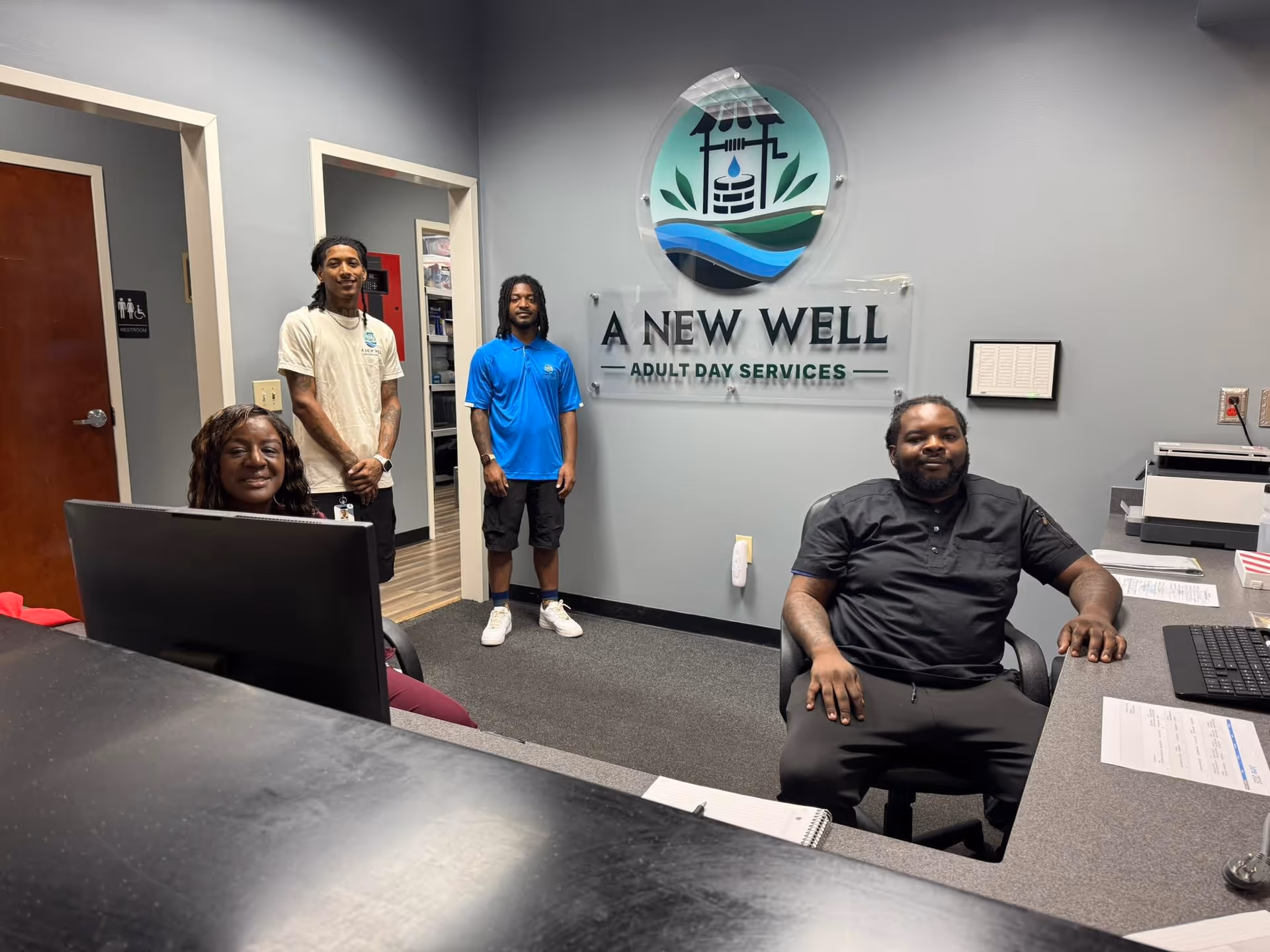 Four staff members stand and sit in a reception area with the A New Well sign.