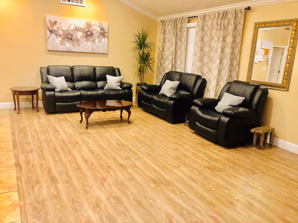 A cozy living room with three black leather recliners and a matching sofa, each with light-colored throw pillows. There is a wooden coffee table in the center and a small side table to the left. A tall green plant is placed in the corner near a window with patterned curtains. A large floral painting hangs on the wall above the sofa, and a gold-framed mirror is mounted on the adjacent wall. The floor is covered with light wood laminate.