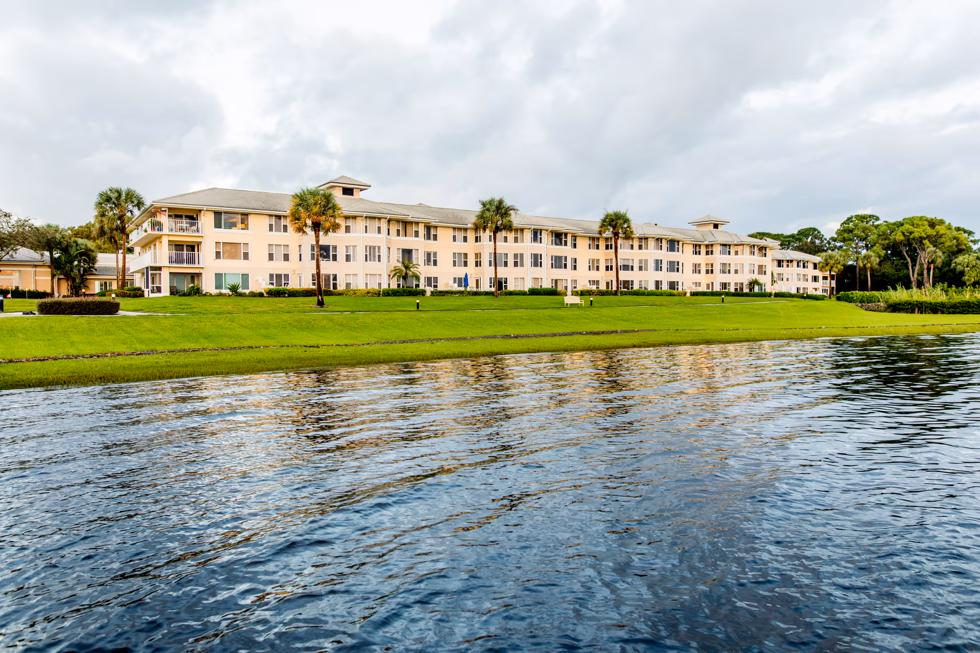 Low-rise beige retirement building with palm trees on a grassy shore reflected in a calm lake.