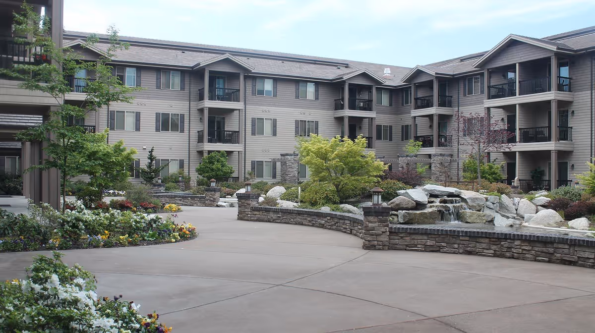 Landscaped courtyard with a stone fountain in front of a multi-story senior living building with balconies.