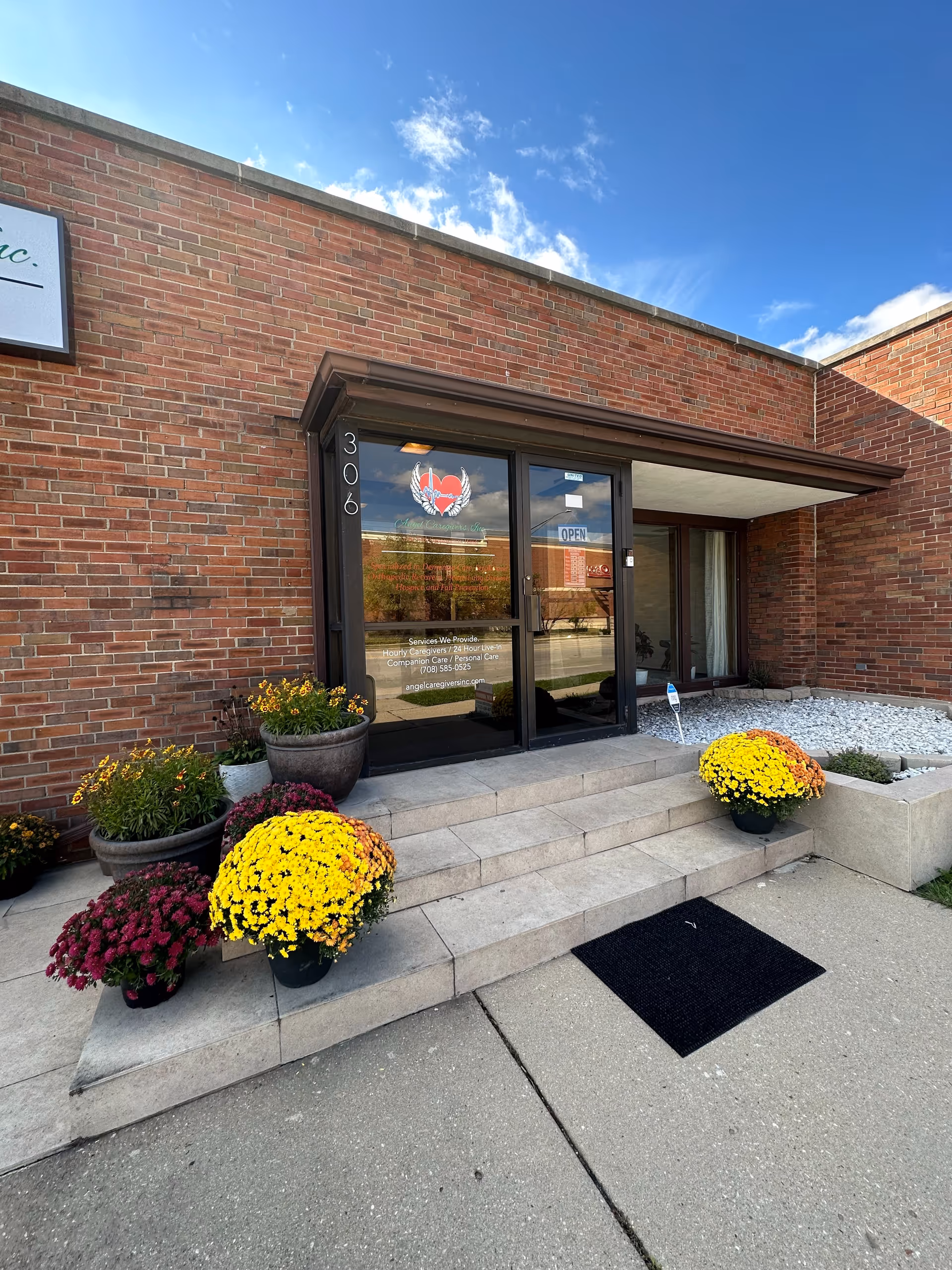 Brick facility entrance with glass doors, steps, and potted flowers outside Angel Caregivers, Inc.