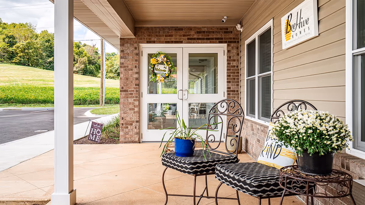 Front entrance of BeeHive Homes of Taylorsville showing double glass doors with a yellow and white floral welcome wreath. There are two black wrought iron chairs with patterned cushions and a small round table holding a potted plant with white flowers. A sign with the BeeHive Homes logo is mounted on the beige siding wall to the right. The area is covered and overlooks a paved driveway and green grassy hill in the background.