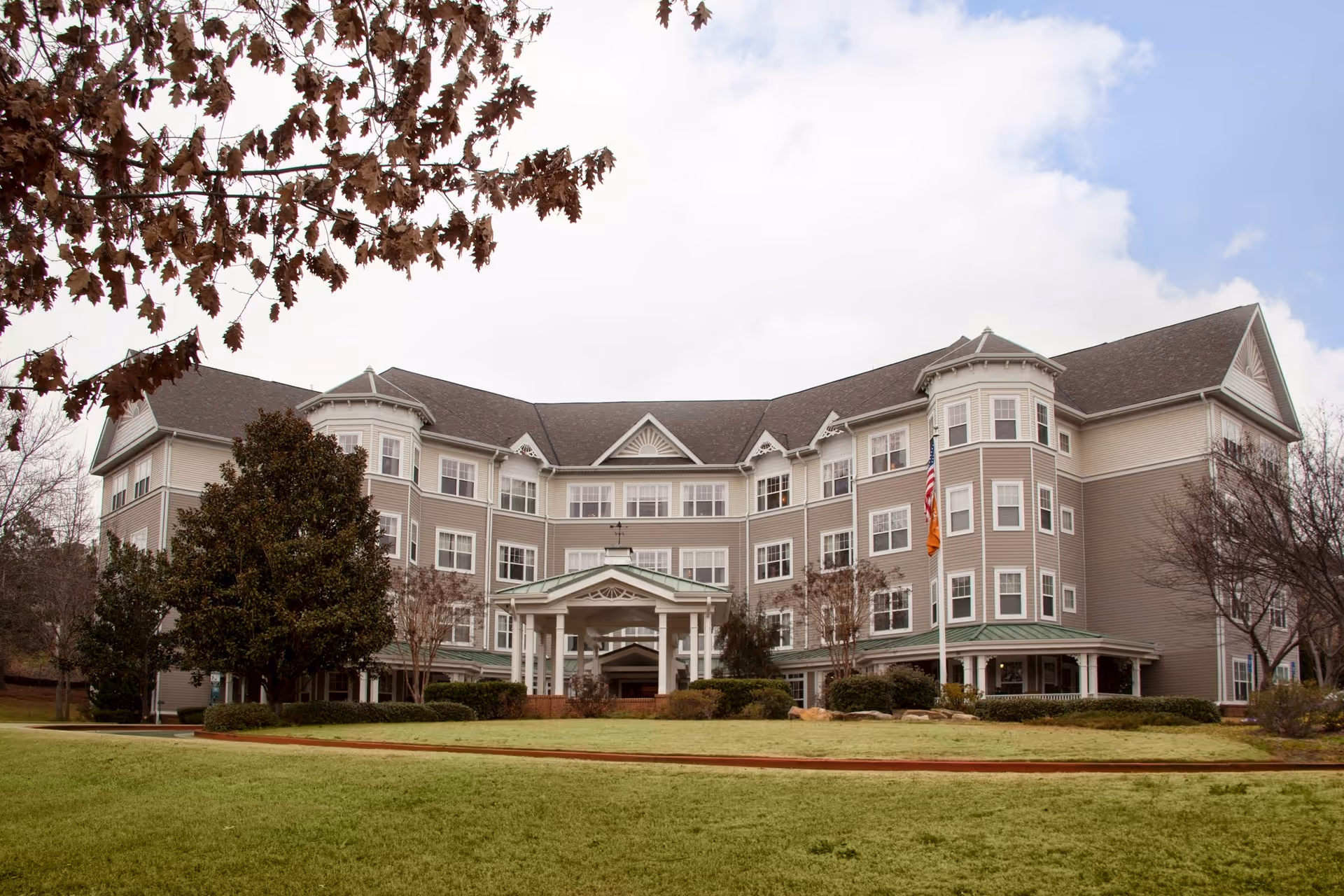 Exterior view of a large, multi-story senior living facility building with beige siding and white trim. The building has multiple windows, a covered entrance with columns, and an American flag on a flagpole in front. The foreground features a well-maintained lawn and some trees with autumn leaves.