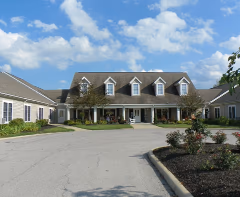 Front exterior of a one-story residence with dormer windows, a covered porch entrance, circular driveway and landscaped beds under a blue sky.