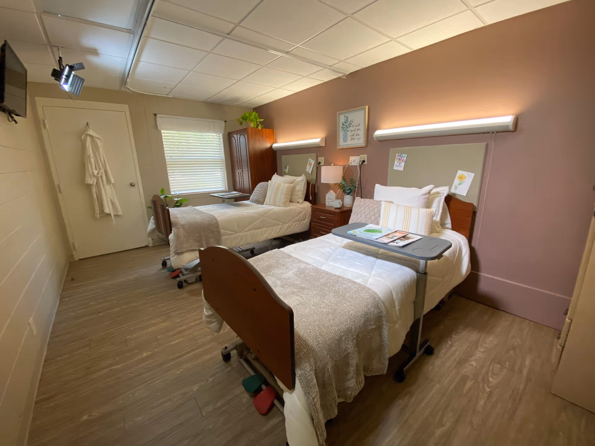 A nursing facility bedroom with two hospital-style beds, each with white bedding and beige blankets. Each bed has a small overbed table with magazines and papers on it. There is a wooden nightstand with a lamp and a plant between the beds. A window with blinds is on the far wall, and a wooden wardrobe is next to it. A white robe hangs on the door, and a flat-screen TV is mounted on the wall to the left.