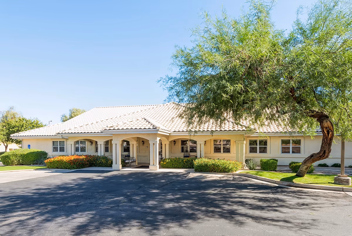 Front exterior view of a single-story building with a tiled roof, beige walls, and a covered entrance supported by columns. There are bushes and a tree in front of the building, with a paved driveway and parking area in the foreground under a clear blue sky.