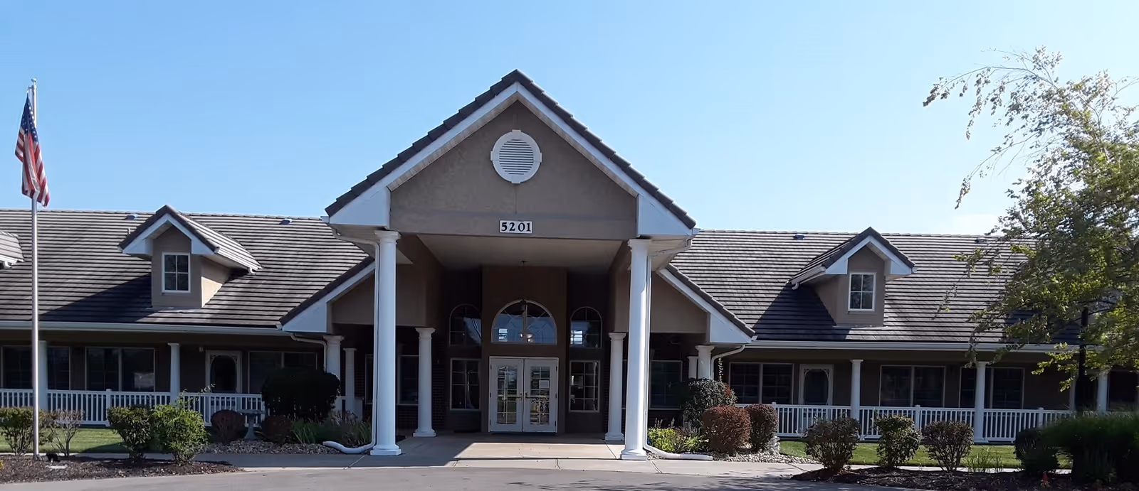 Front entrance of a single-story senior living building with white columns, a covered portico, and landscaped shrubs under a clear sky.
