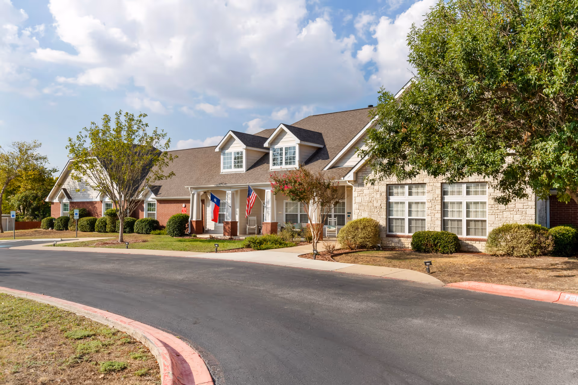 Exterior view of a senior living facility building with a combination of brick and stone facade, two dormer windows on the roof, and two flags (Texas state flag and American flag) displayed near the entrance. The building is surrounded by neatly trimmed bushes, trees, and a paved driveway with a red curb.