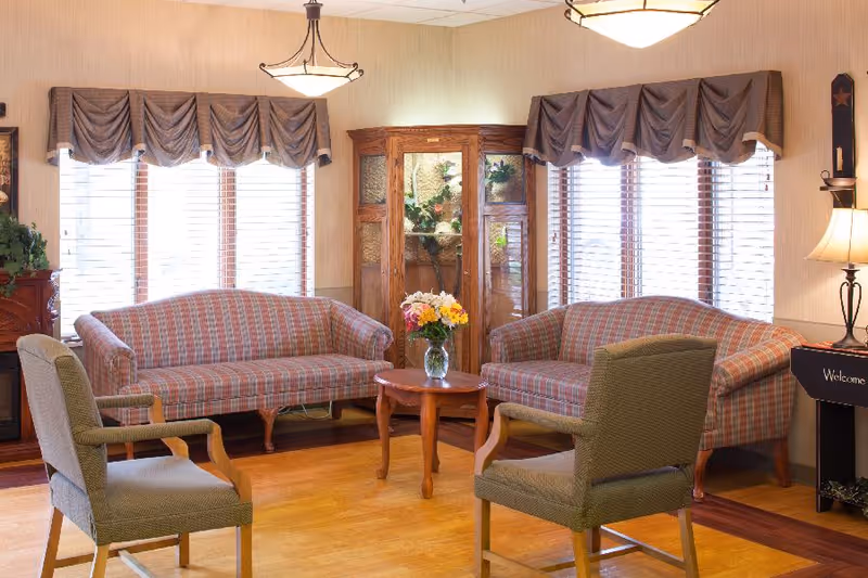 A cozy living room area with two plaid sofas facing each other and two green armchairs. A small wooden coffee table with a vase of colorful flowers is in the center. The room has large windows with blinds and valances, a wooden display cabinet with glass doors, and warm lighting from ceiling fixtures and a table lamp.