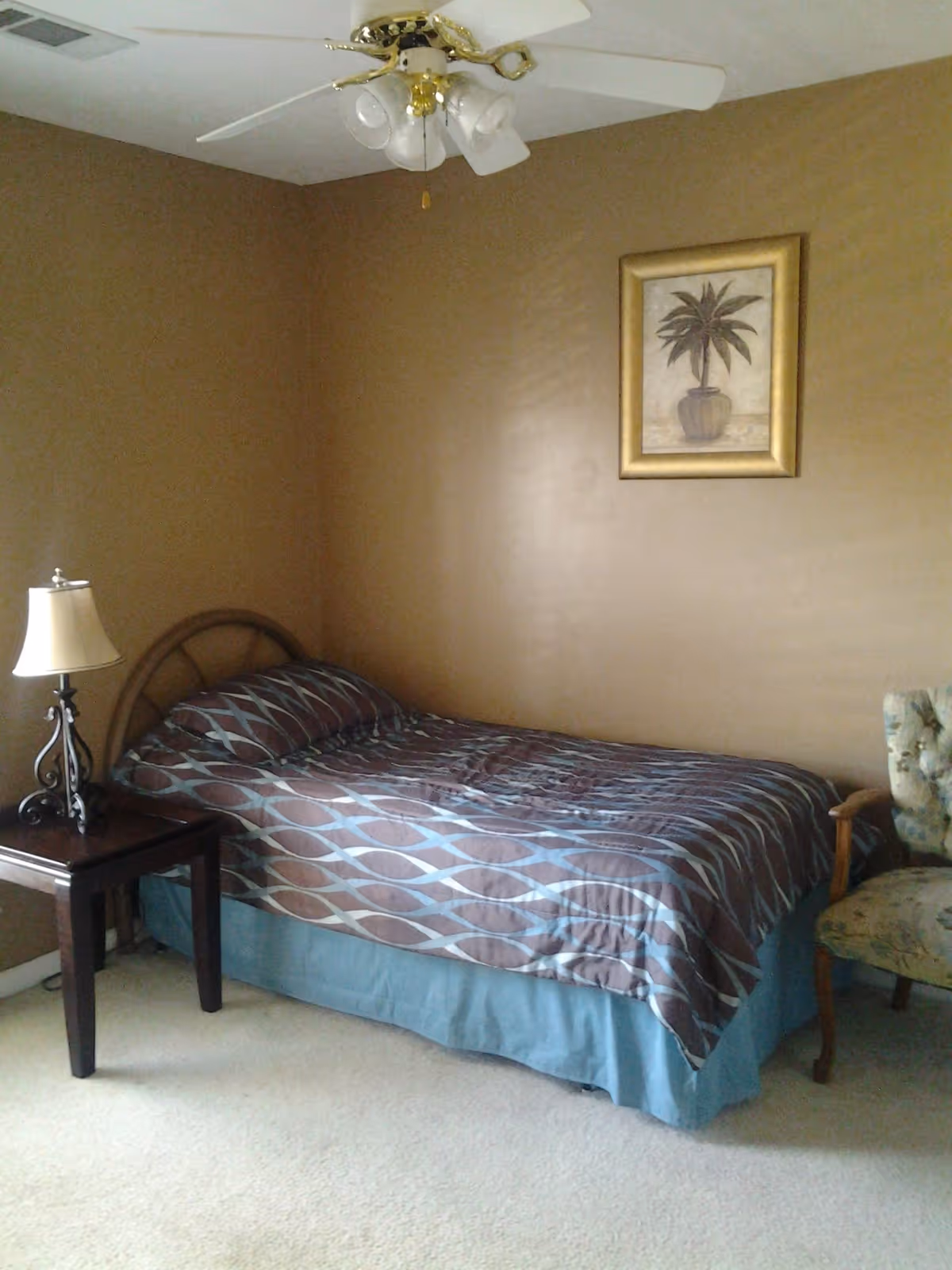 A furnished bedroom with a bed, nightstand, lamp, ceiling fan, and palm painting.