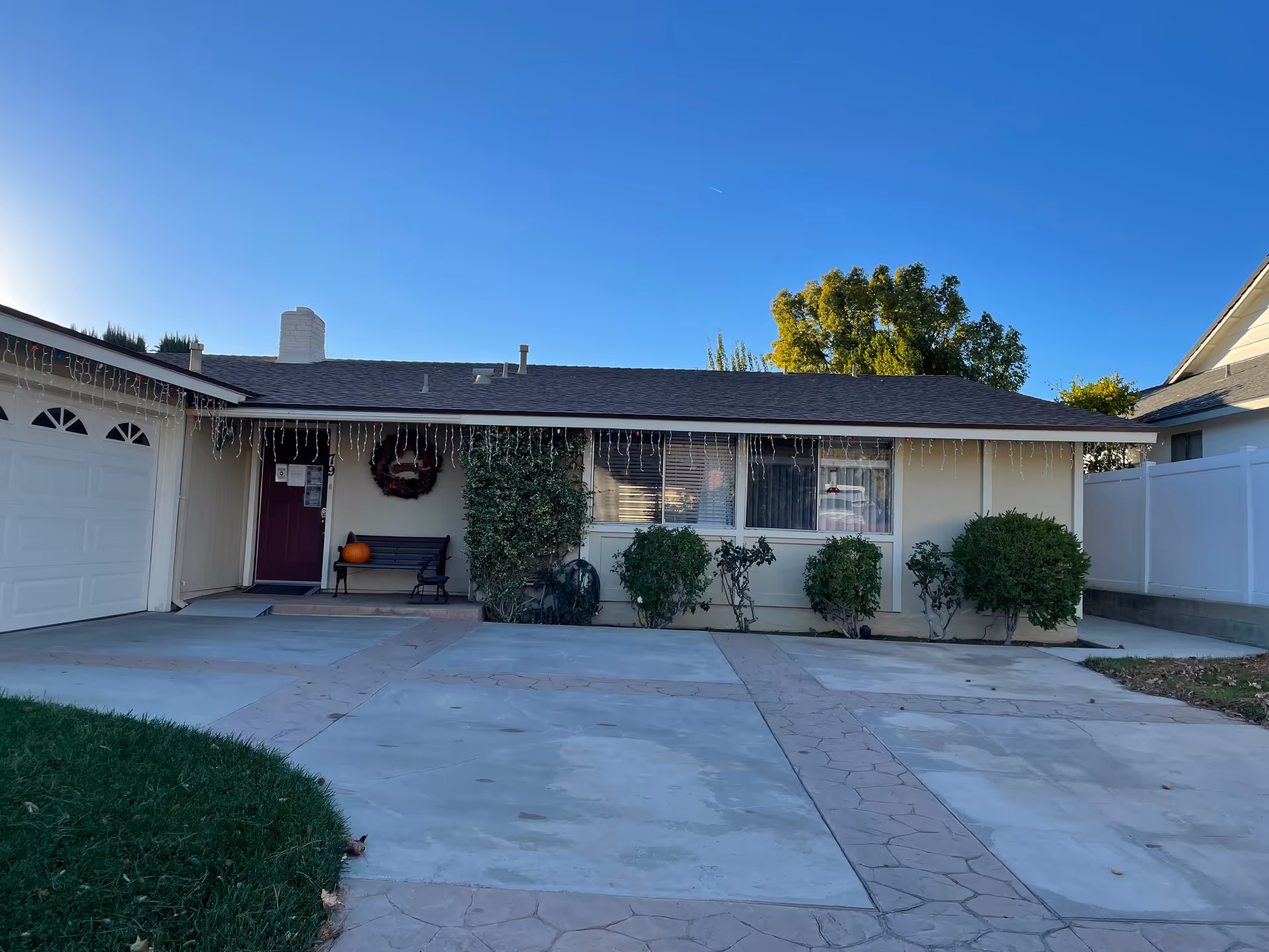 Single-story residential building with a garage on the left, a front porch with a bench and a pumpkin, bushes along the front wall, and icicle-style holiday lights hanging from the roof edge under a clear blue sky.