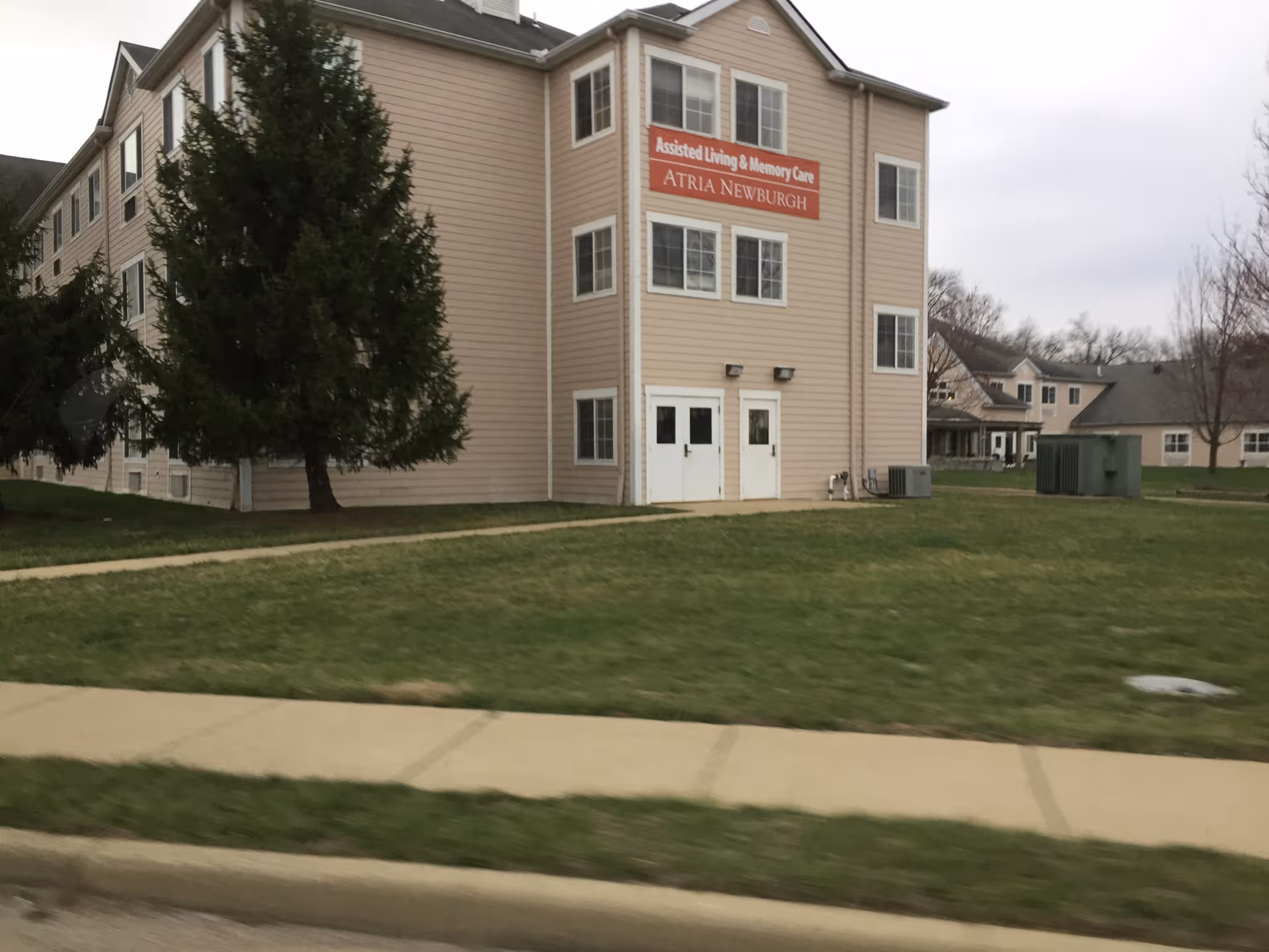 Two-story beige senior living building with sign, trees, and a grassy lawn.