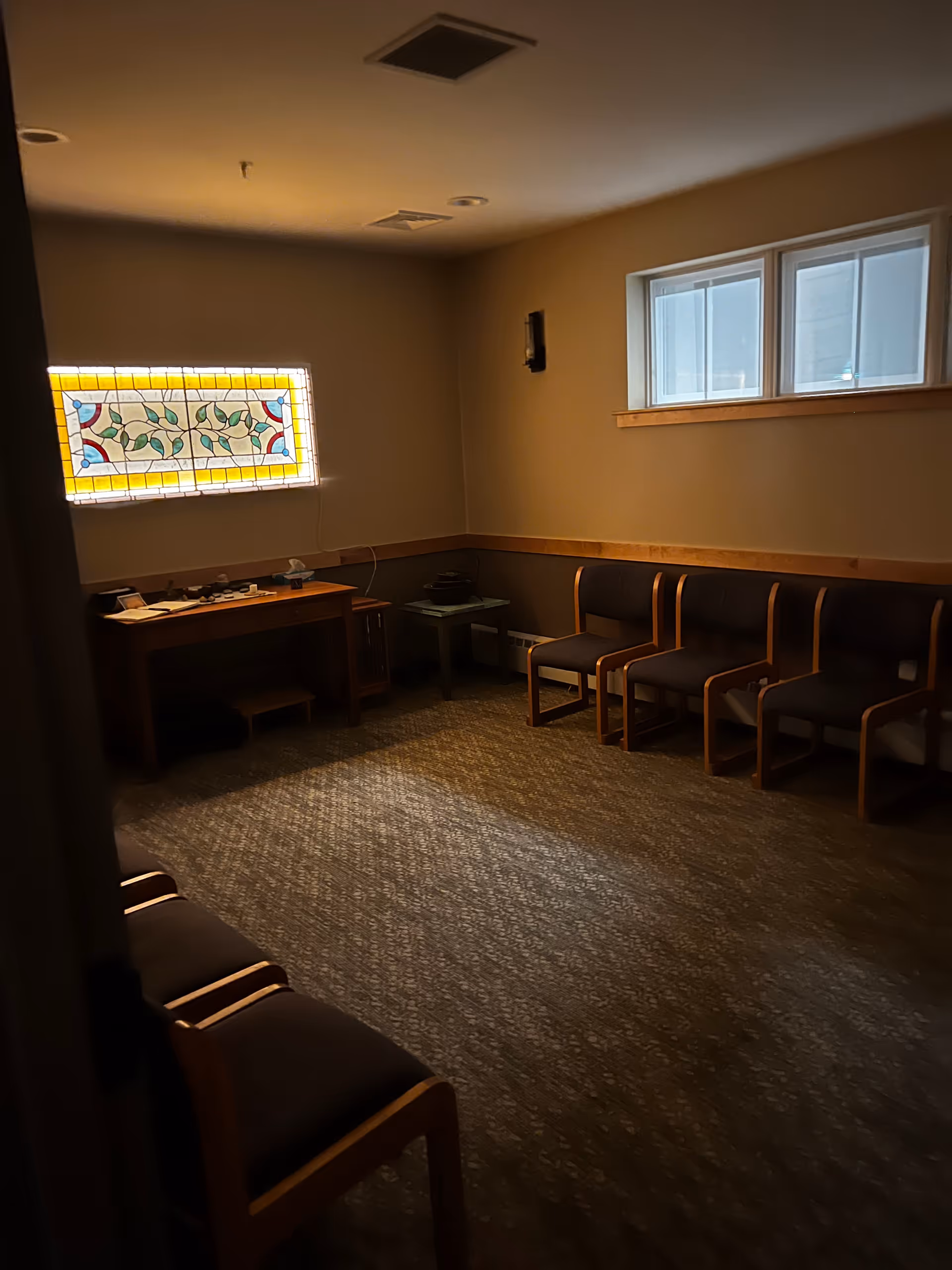 Dim interior room with a row of chairs, a small desk, and a stained glass window.
